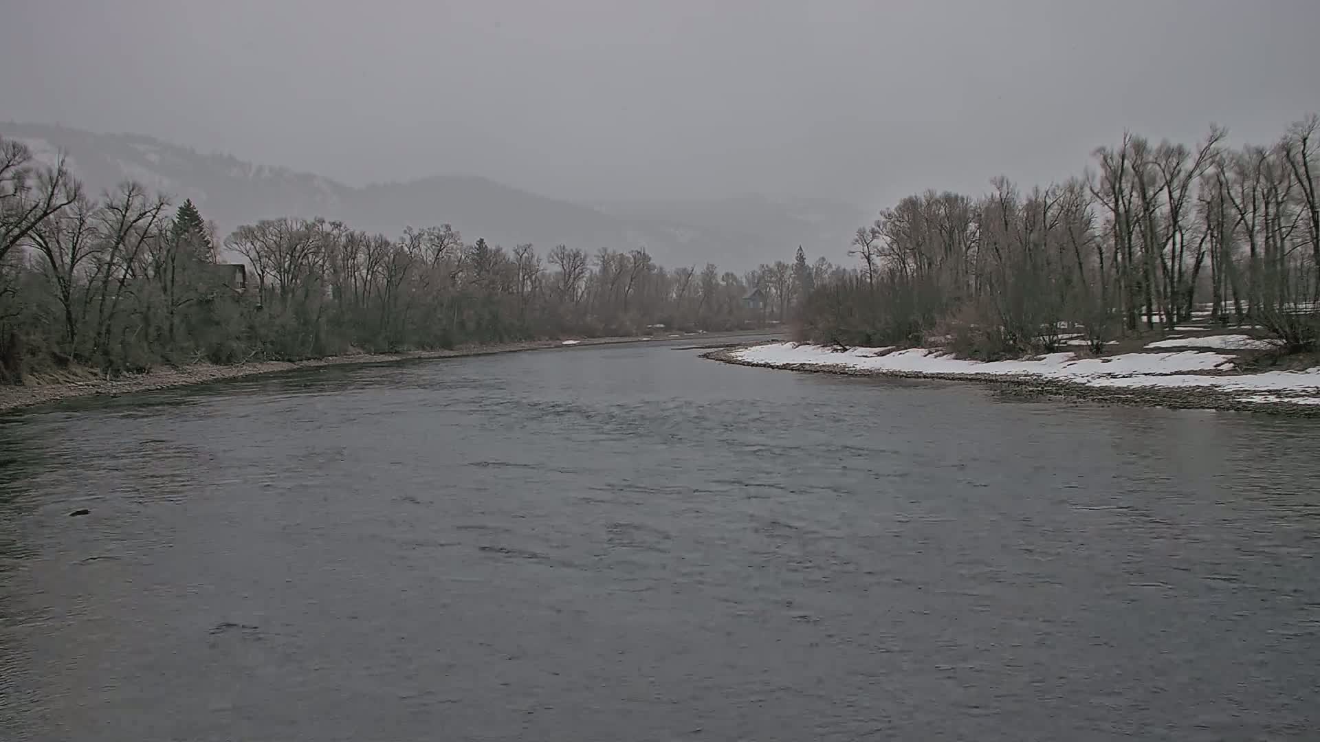 A wide, gray river bends gently past snow-dusted banks lined with bare, winter trees under a hazy, overcast sky.