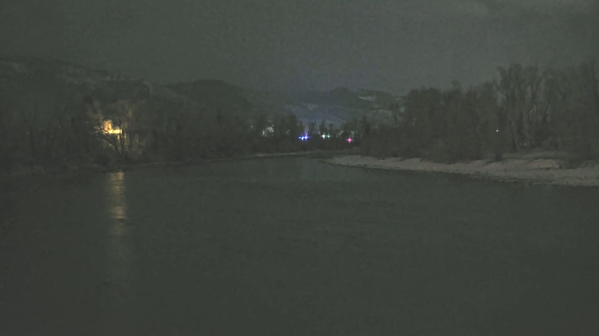 A dark, calm river flows under a nighttime sky past snow-covered banks lined with dark trees and distant hills.