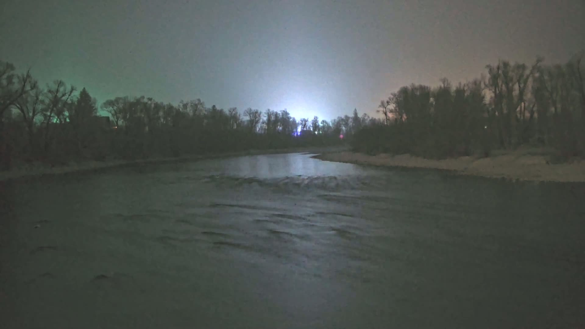 A dark, murky river flows under a muted, nighttime sky past a snow-dusted shoreline lined with leafless trees.