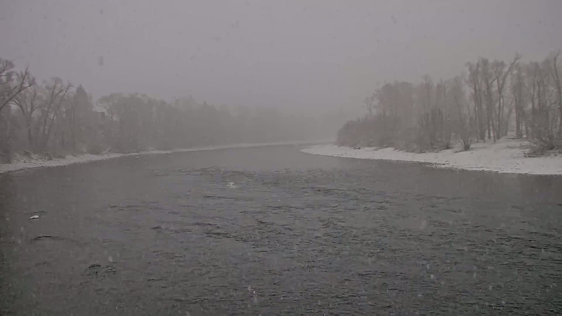 A snow-covered river flows between banks lined with snow-laden trees during a heavy snowfall.