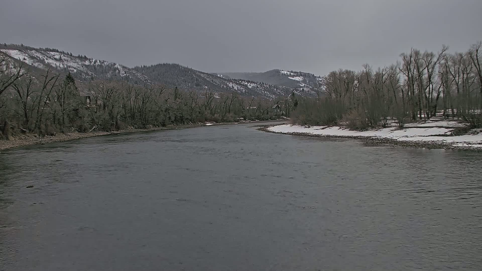 A wide, grey river flows through a valley under a cloudy sky, with snow-dusted hills in the background and leafless trees lining the riverbanks.