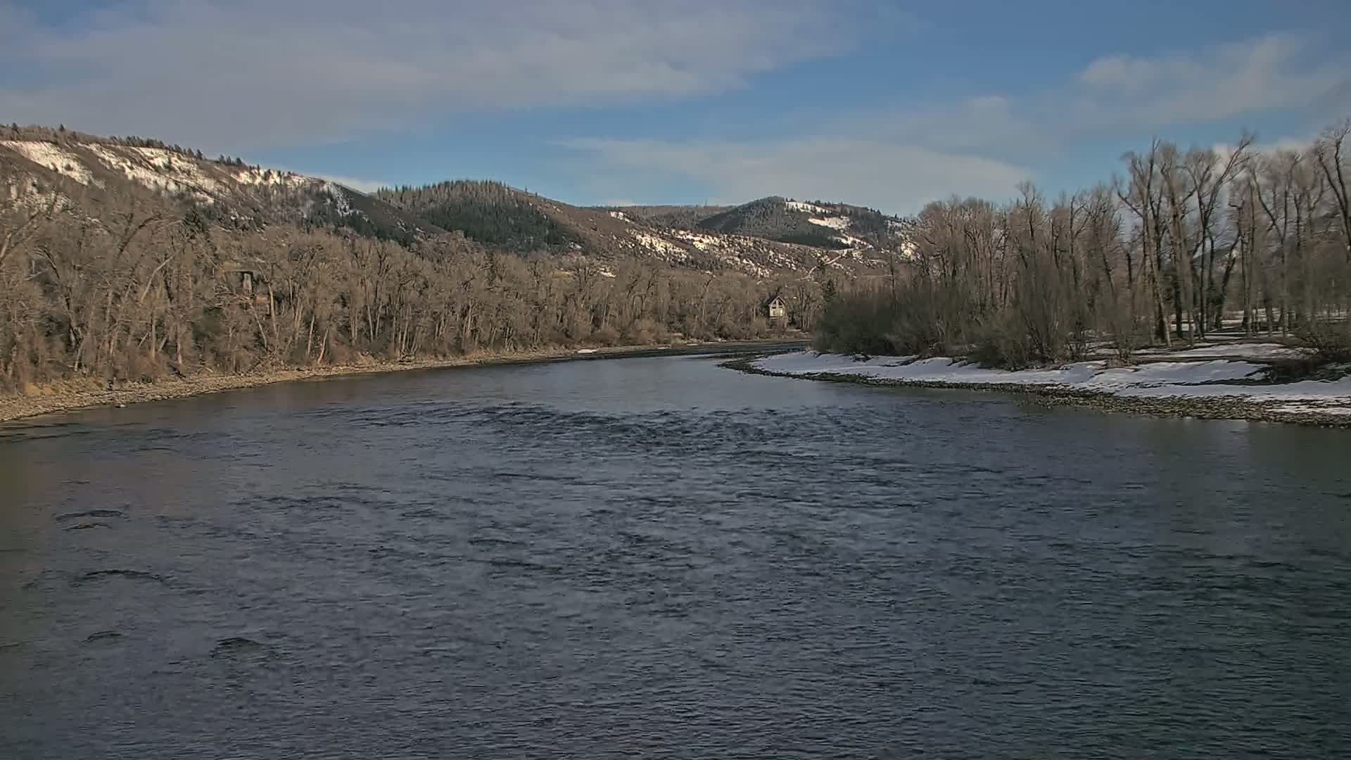 A wide, dark river flows through a valley with snow-dusted banks and leafless trees under a mostly sunny sky.