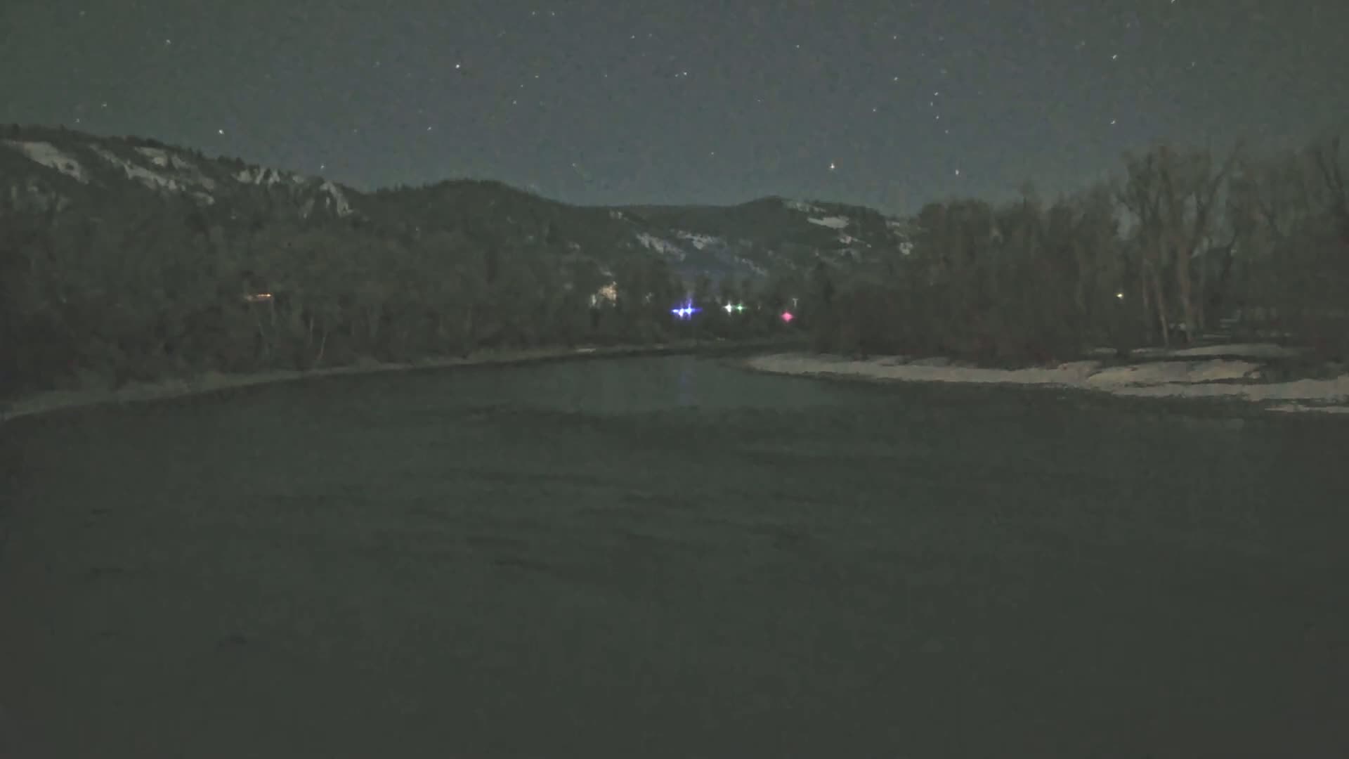 A dark river flows under a starry night sky past snow-covered banks and dark, leafless trees bordering hills in the distance.