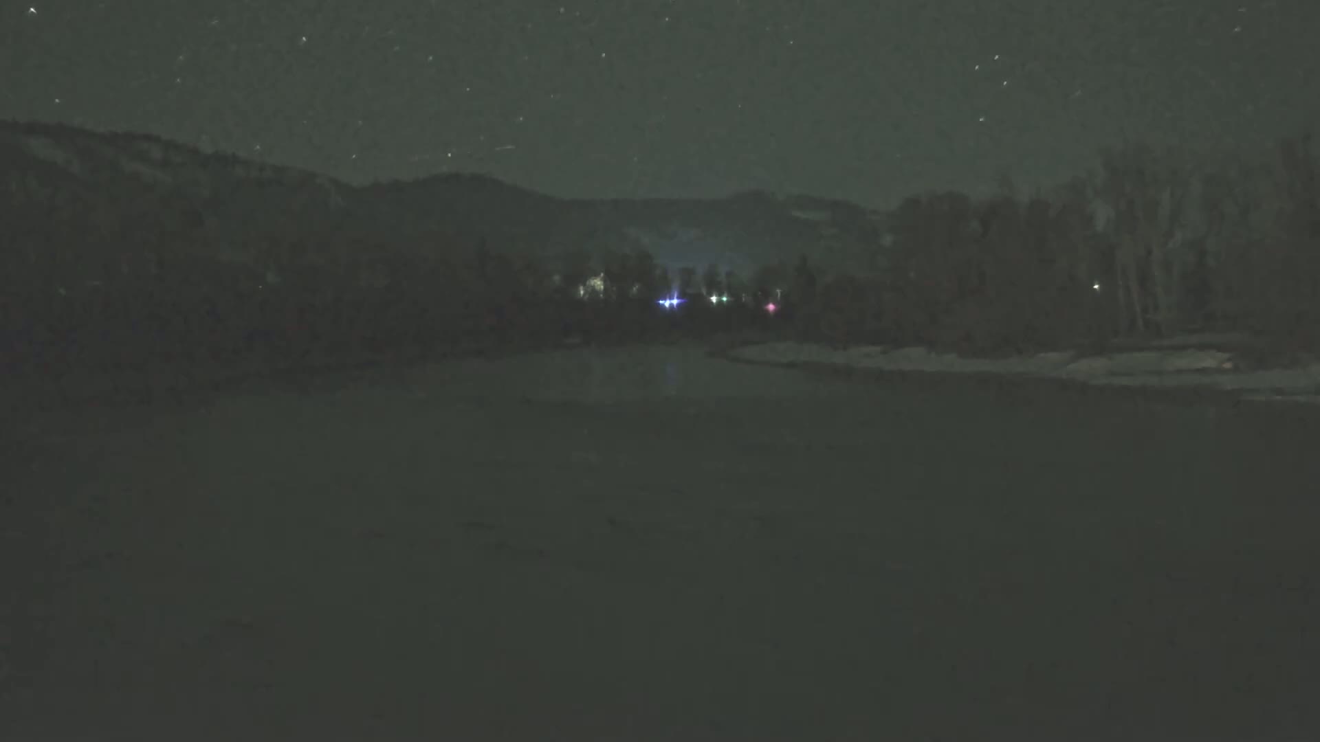 Under a starry night sky, a dark body of water lies before a snow-covered bank and a line of trees silhouetted against a mountain range.