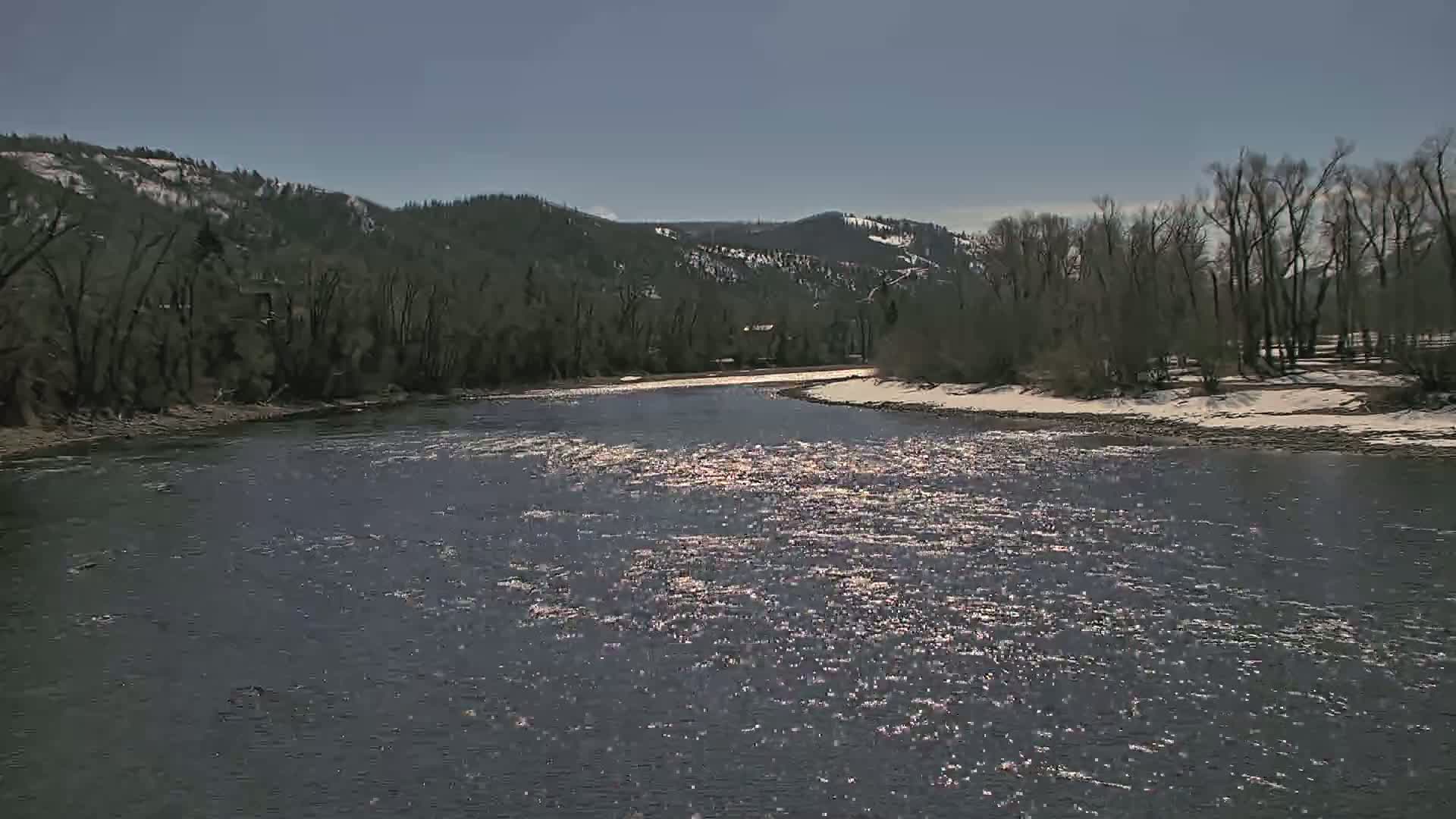 A sparkling river flows under a clear blue sky past snow-dusted banks lined with leafless trees and distant snow-capped mountains.