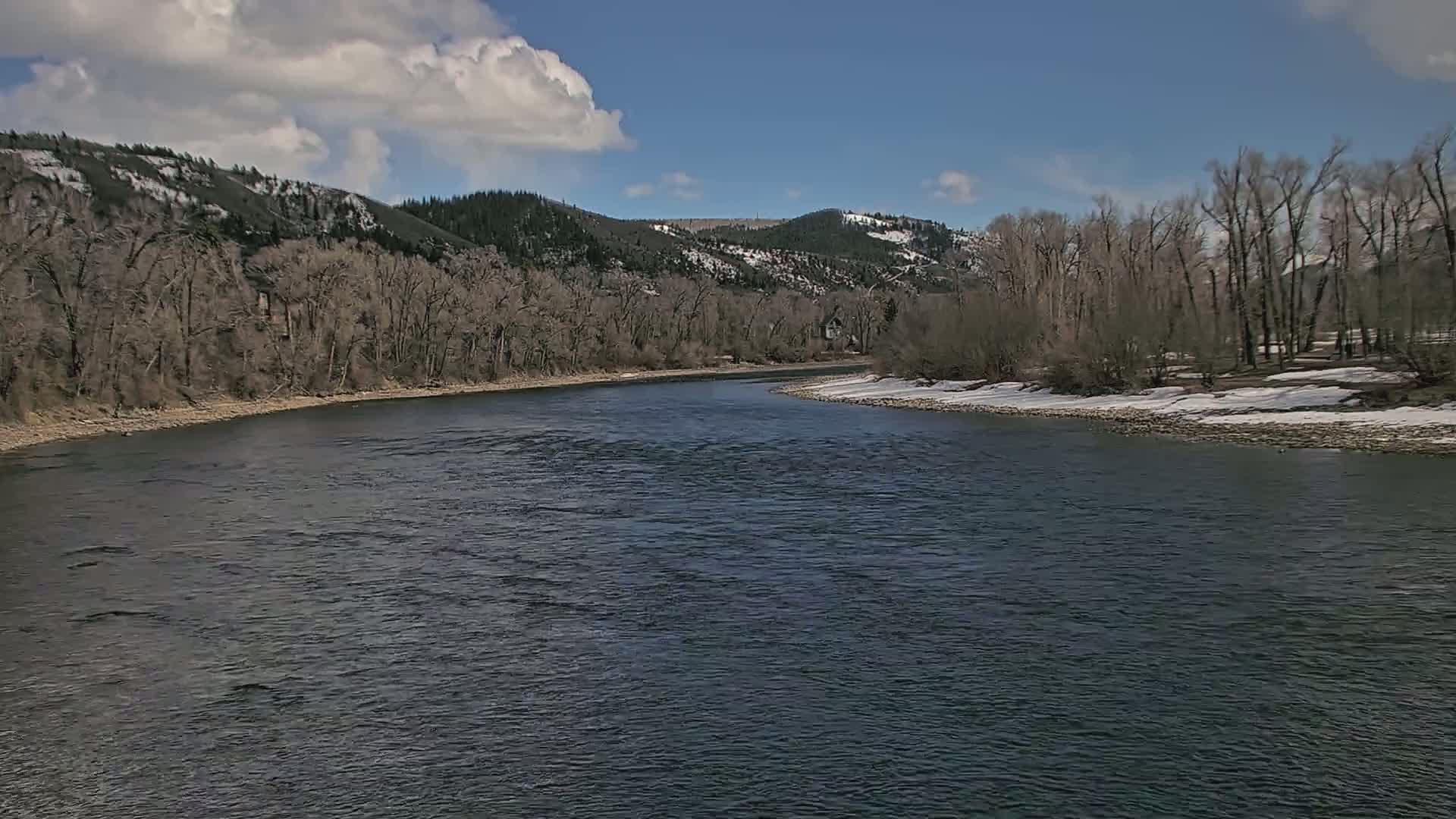 A dark river flows through a valley under a partly cloudy sky, with bare trees lining the riverbanks and snow patches on the ground.