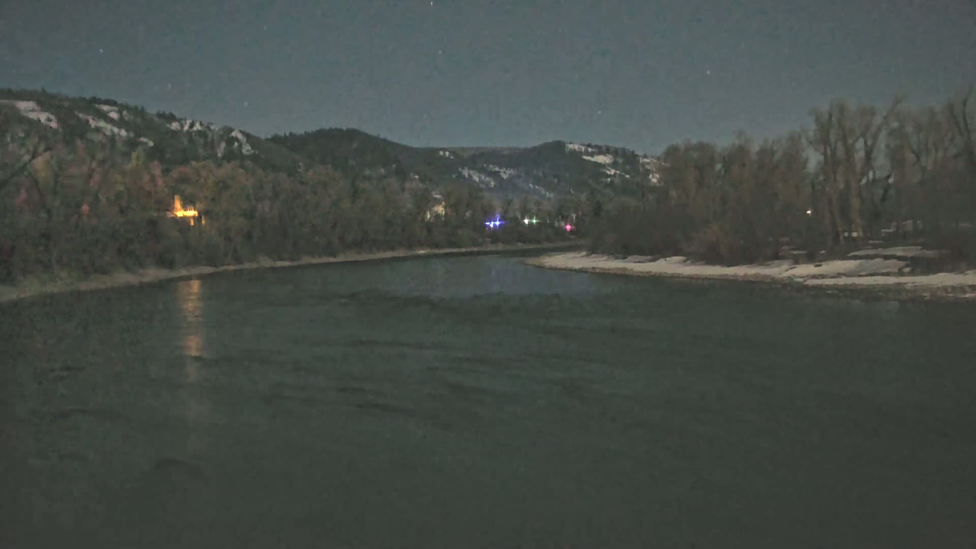 A dark, moonlit night scene shows a river flowing past snow-dusted banks lined with trees and hills under a starry sky.