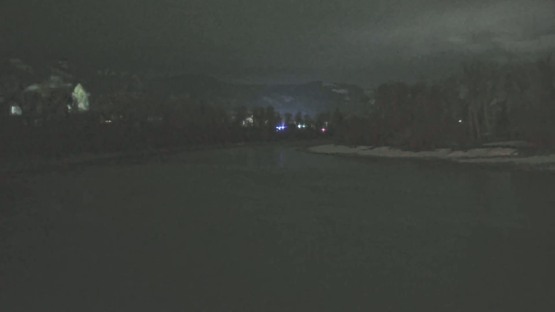A dark body of water is seen at night under a cloudy sky, with dimly lit trees and distant lights on the far shore.