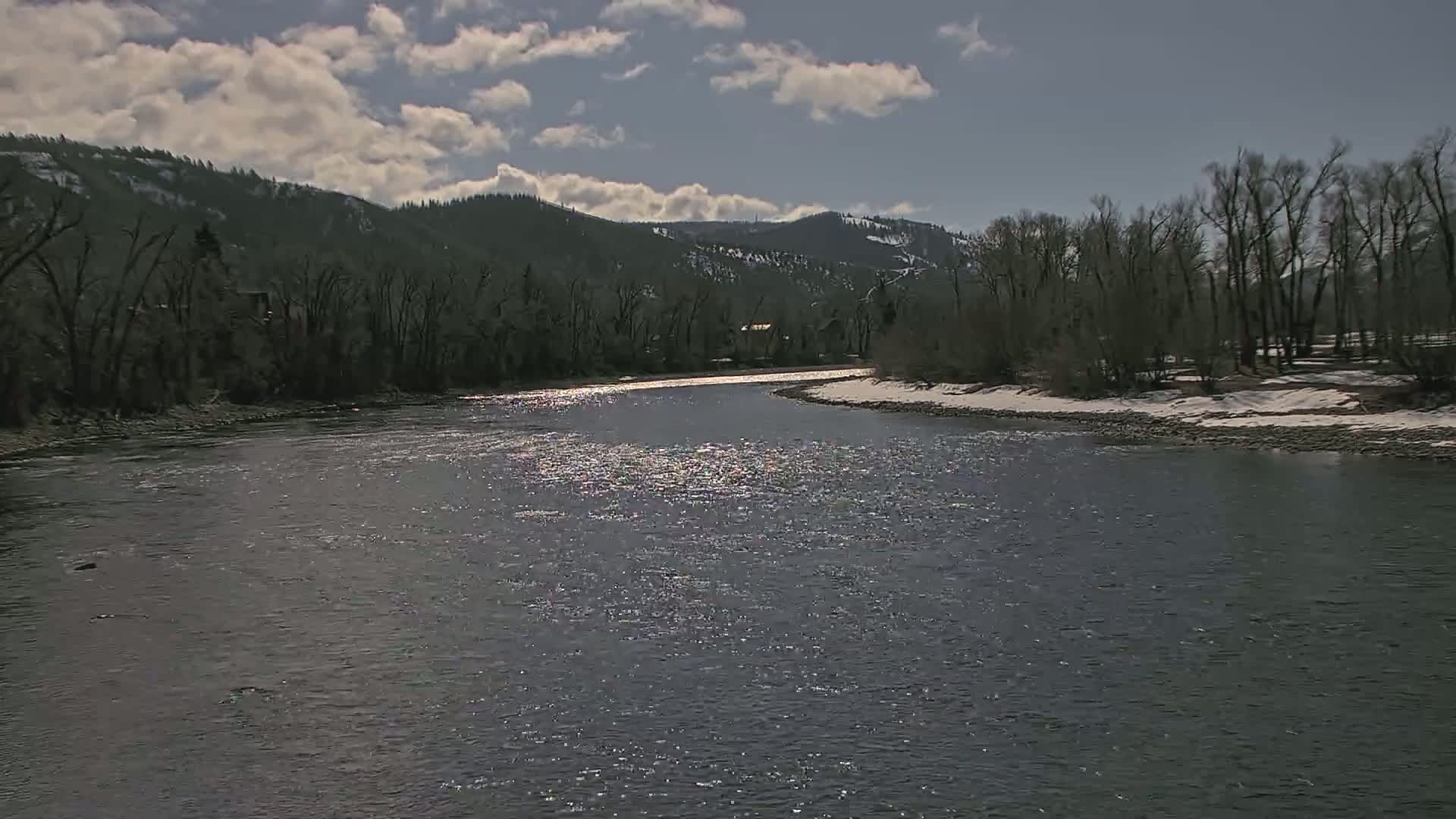 A partly sunny day shows a wide river reflecting sunlight, winding past a snow-dusted riverbank lined with bare trees and a mountainous background.