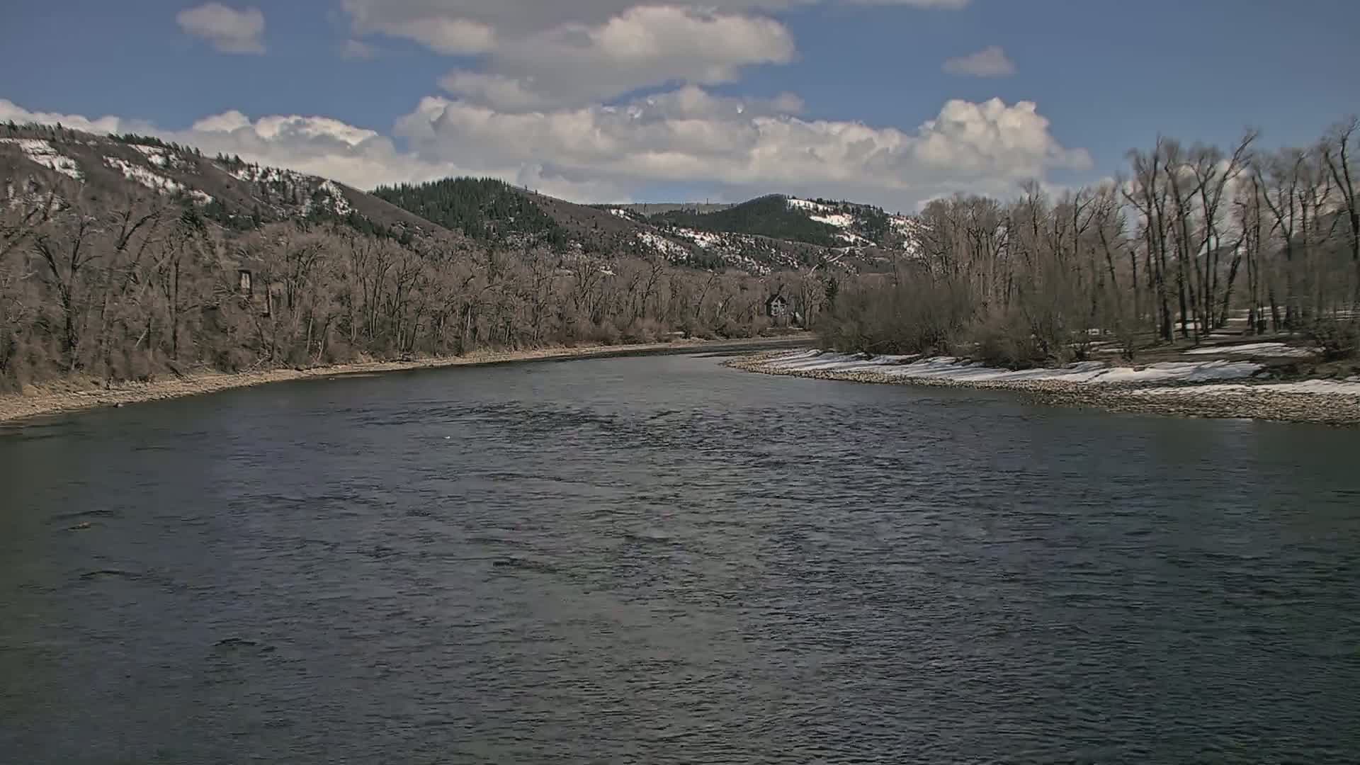 A wide, dark river flows past a snow-dusted shoreline lined with bare trees and hills under a partly cloudy sky.