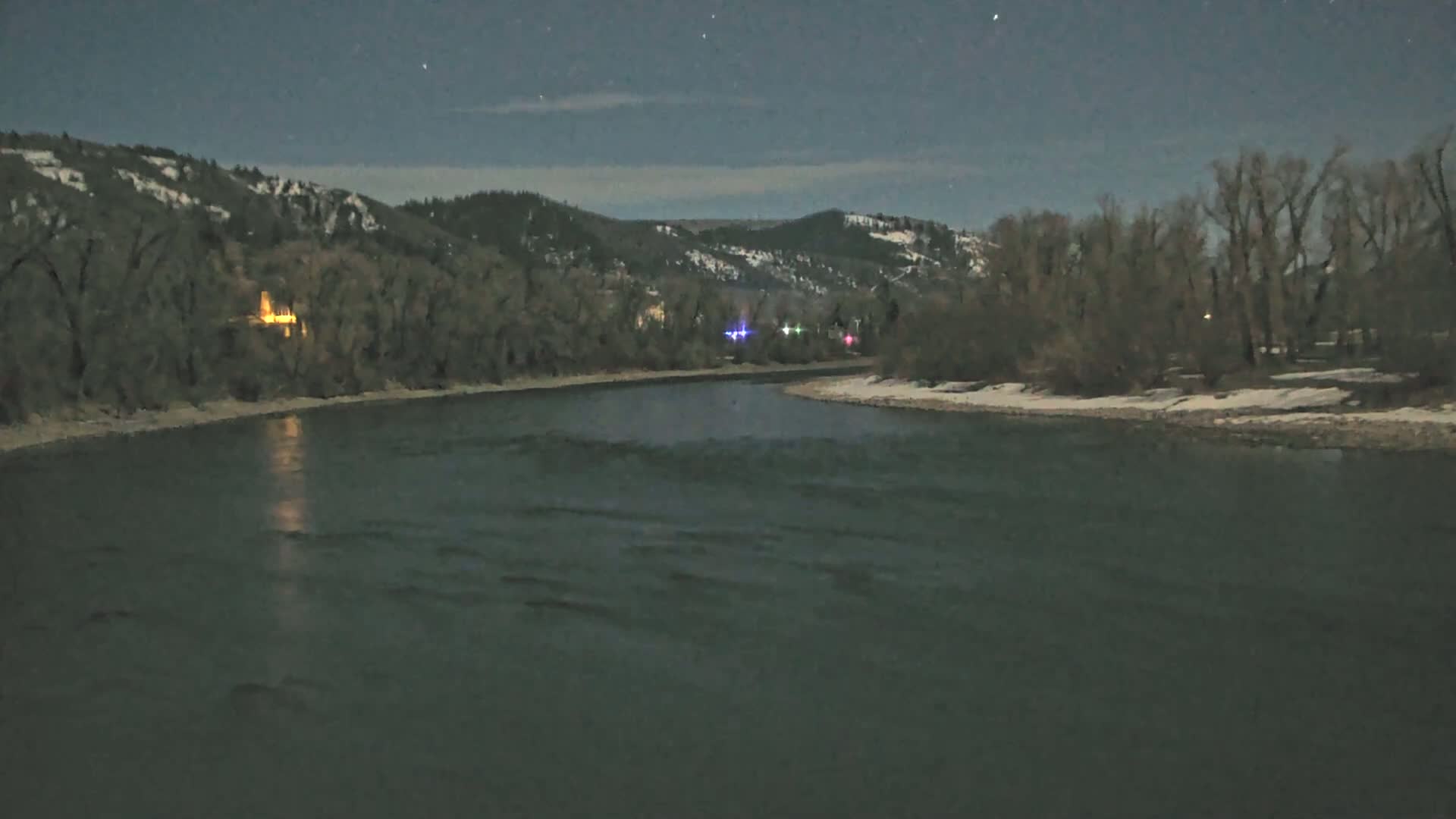 A moonlit river bends around a snow-dusted riverbank lined with bare trees under a clear night sky.