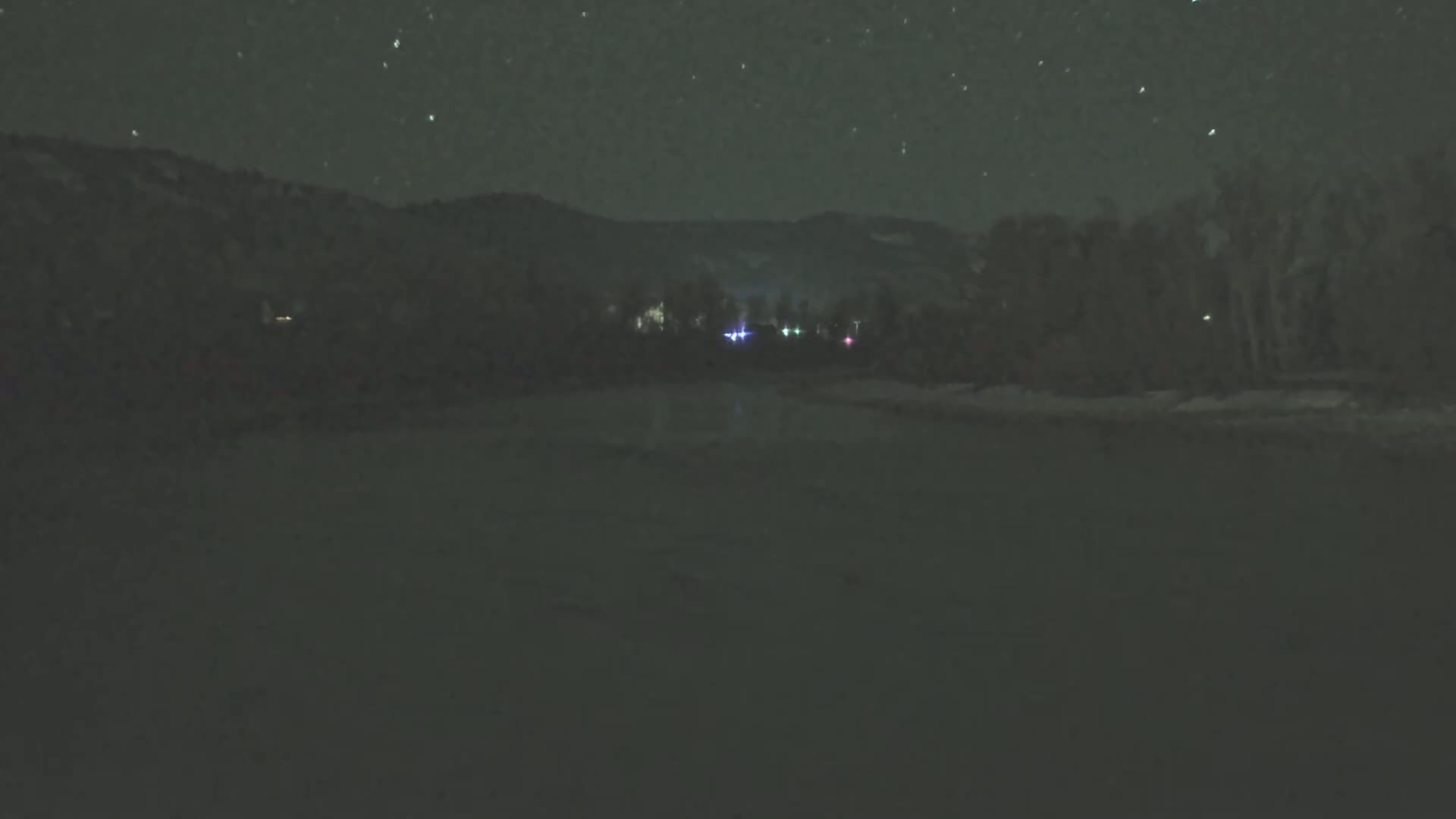 A nighttime, snowy landscape shows a dark, snow-covered foreground with a line of trees and distant mountains under a starry sky.