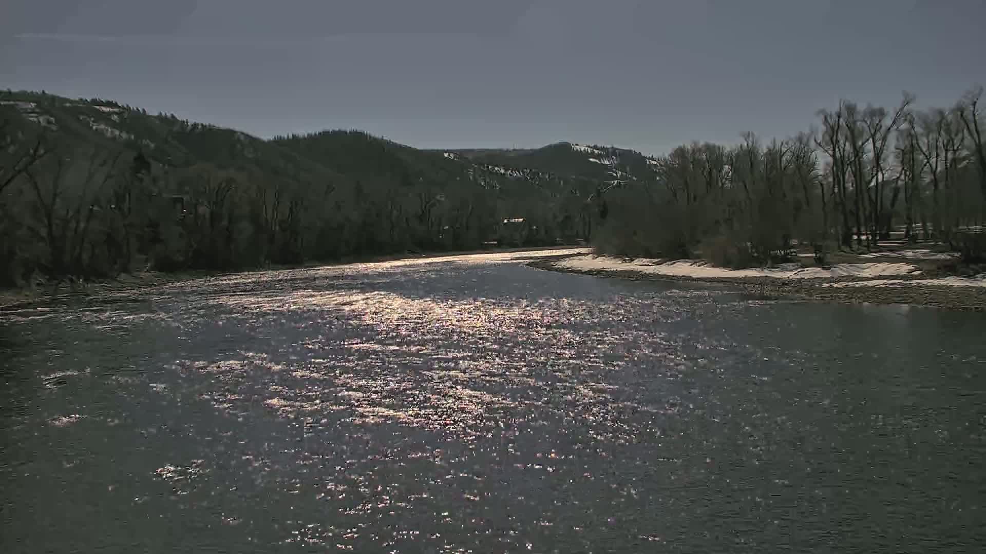 A wide, sunlit river flows past snow-dusted banks and leafless trees under a clear sky.