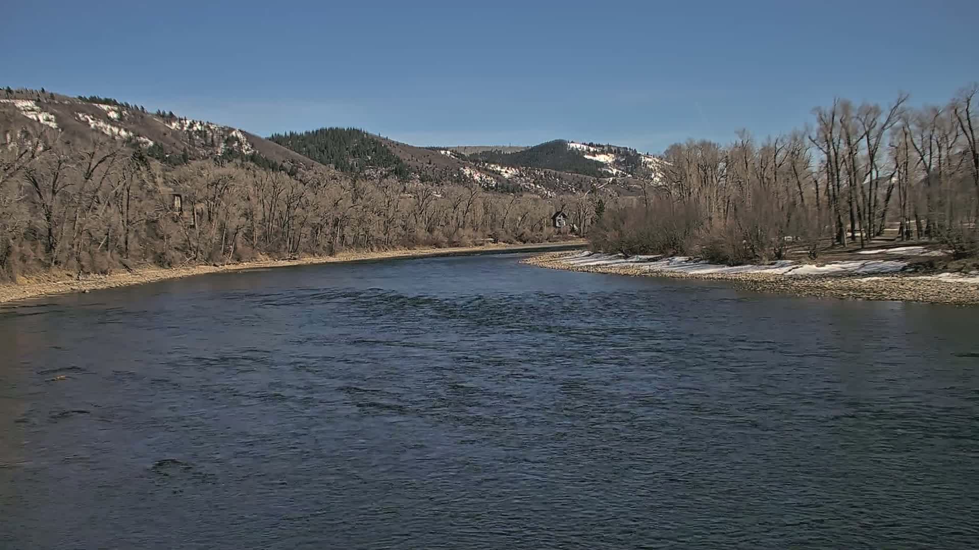 A wide, dark river flows through a valley under a clear blue sky, past barren trees and snow-dusted hillsides.