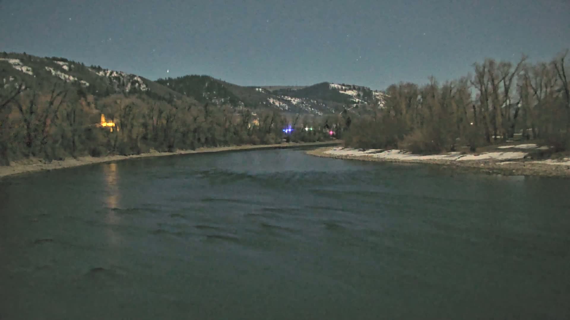 A dark river flows under a night sky past snow-dusted banks lined with leafless trees and hills in the background.