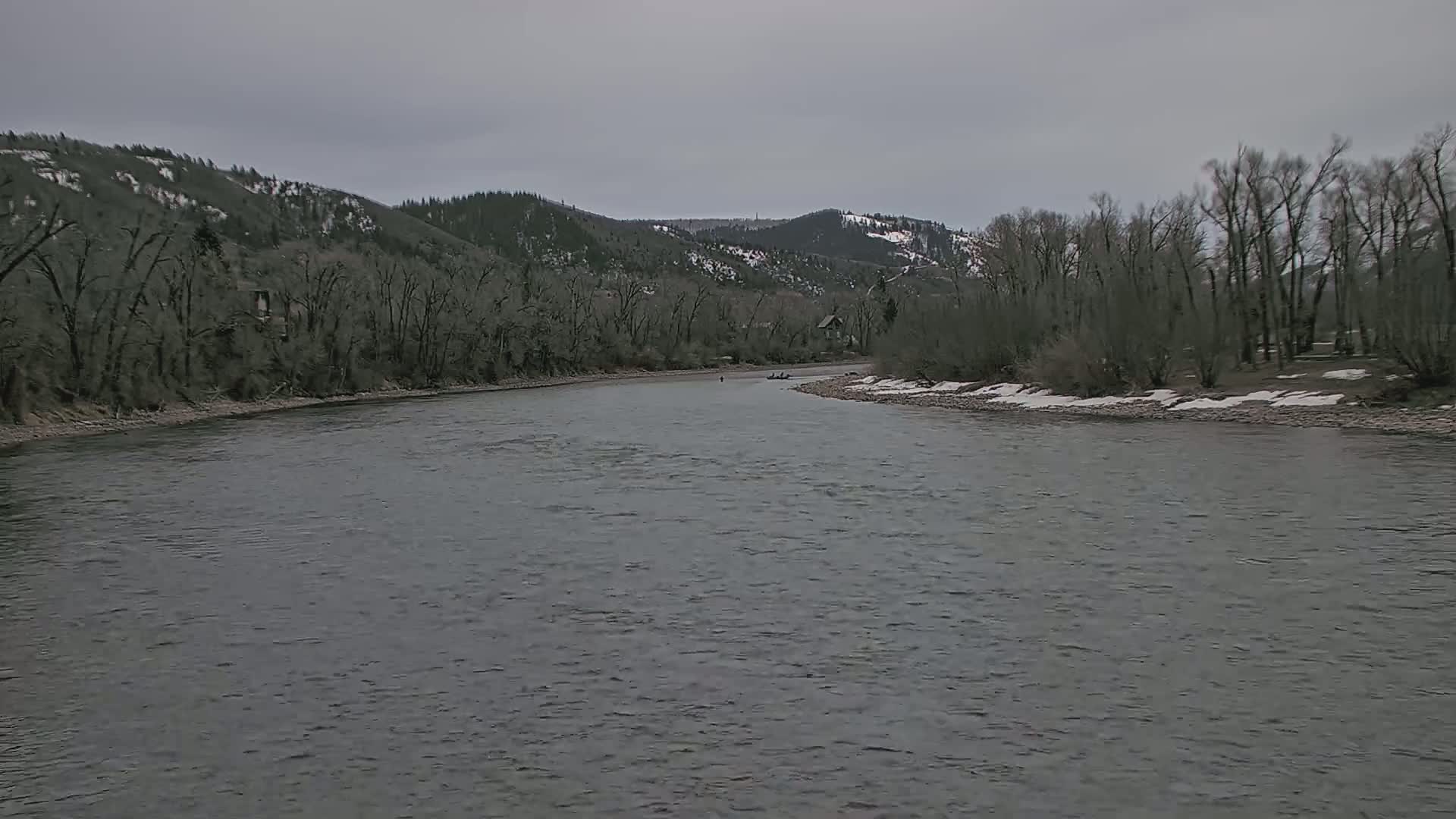 A wide, gray river flows through a valley of bare, dark trees under an overcast sky, with patches of snow visible on the riverbanks and distant hills.