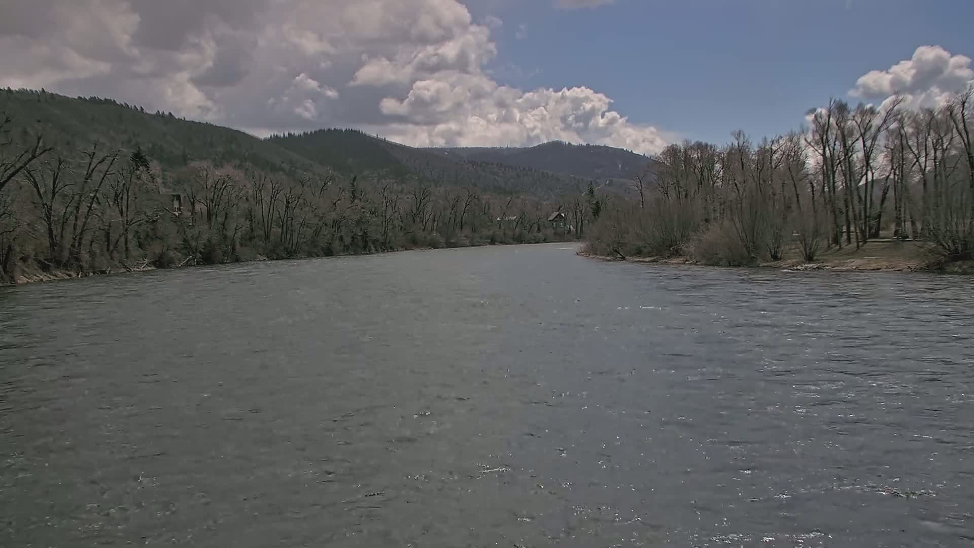 A wide, grey river flows under a partly cloudy sky past bare trees and forested hills.