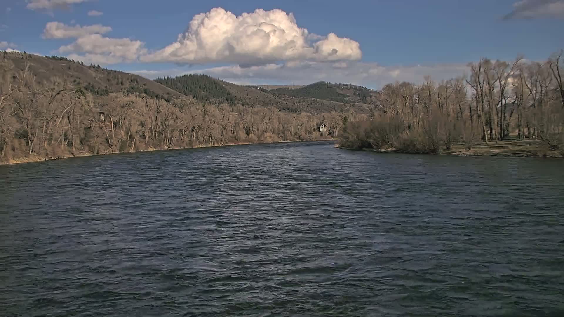 A wide, dark river flows beneath a partly cloudy blue sky, past barren winter trees lining its banks and hills in the distance.