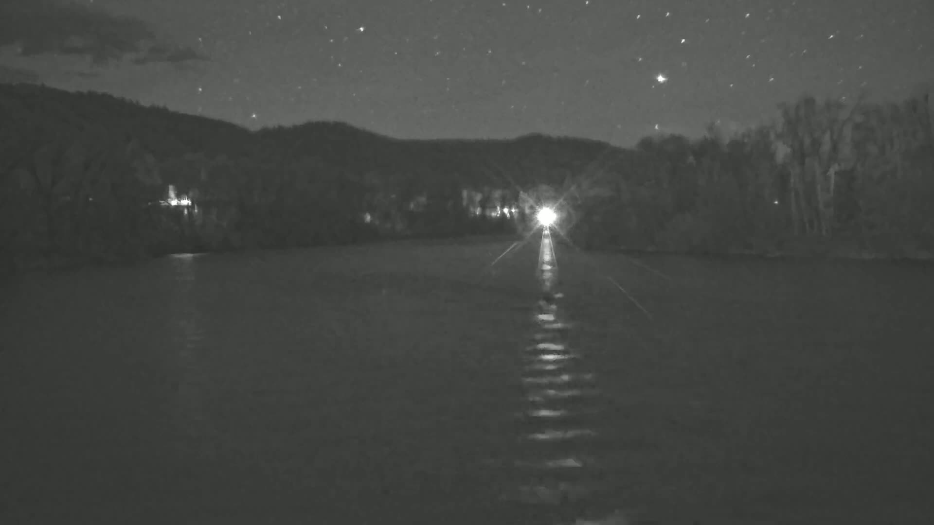 A bright light shines on a dark river at night under a starry sky, with a treeline in the background.