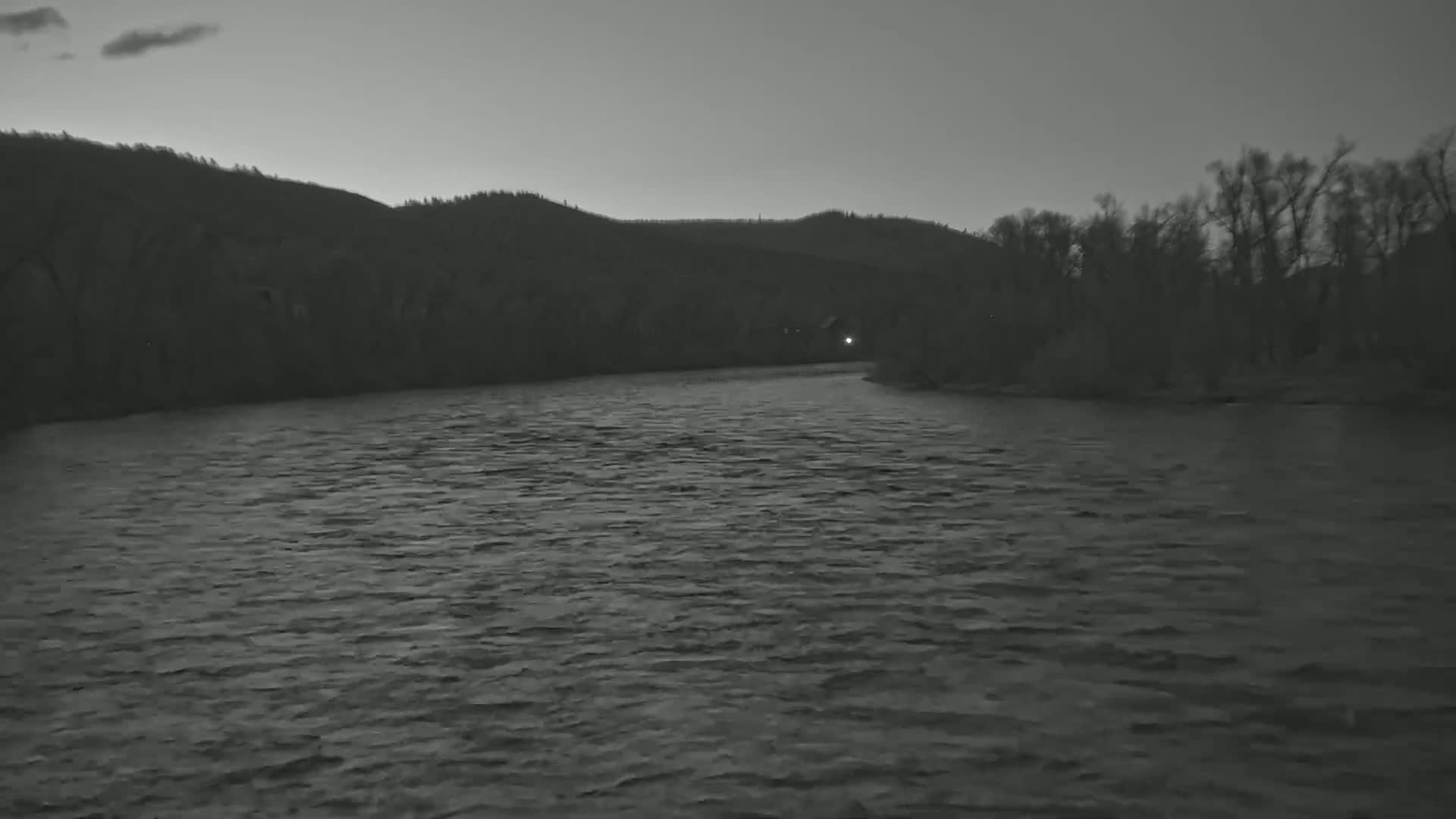 A wide, dark river flows under a dusky sky toward a line of dark trees and hills.