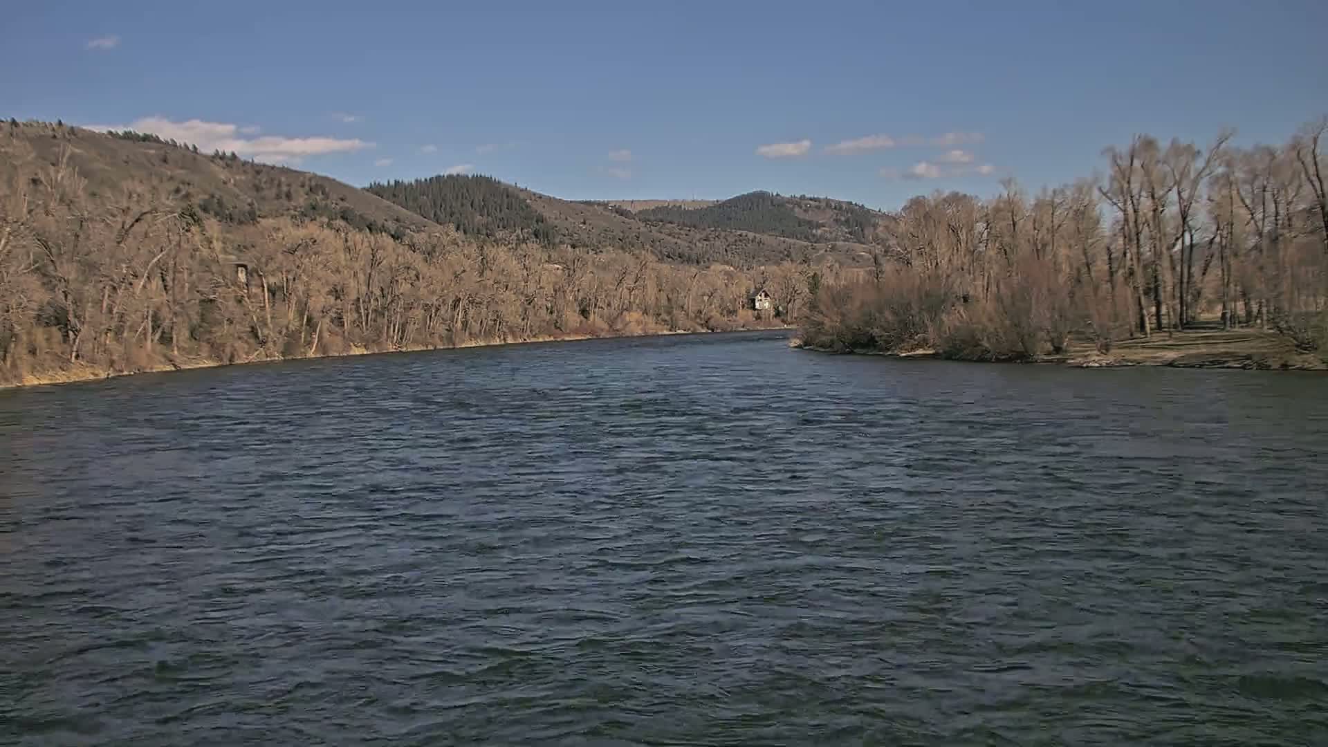 A wide, dark river flows under a clear blue sky past bare trees and hills.