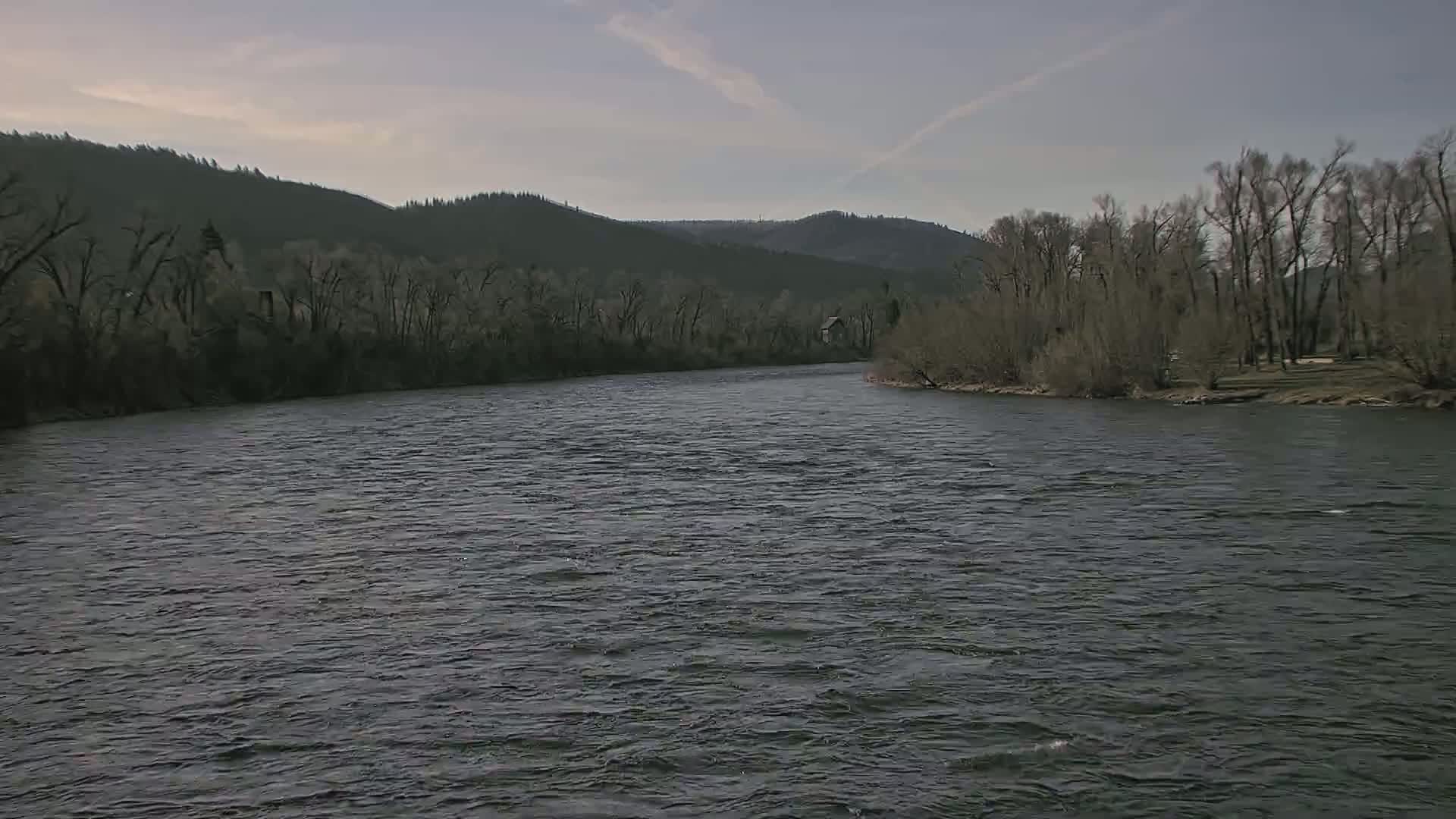 A wide, dark river flows gently under a cloudy sky, bordered by bare winter trees and distant, dark green hills.