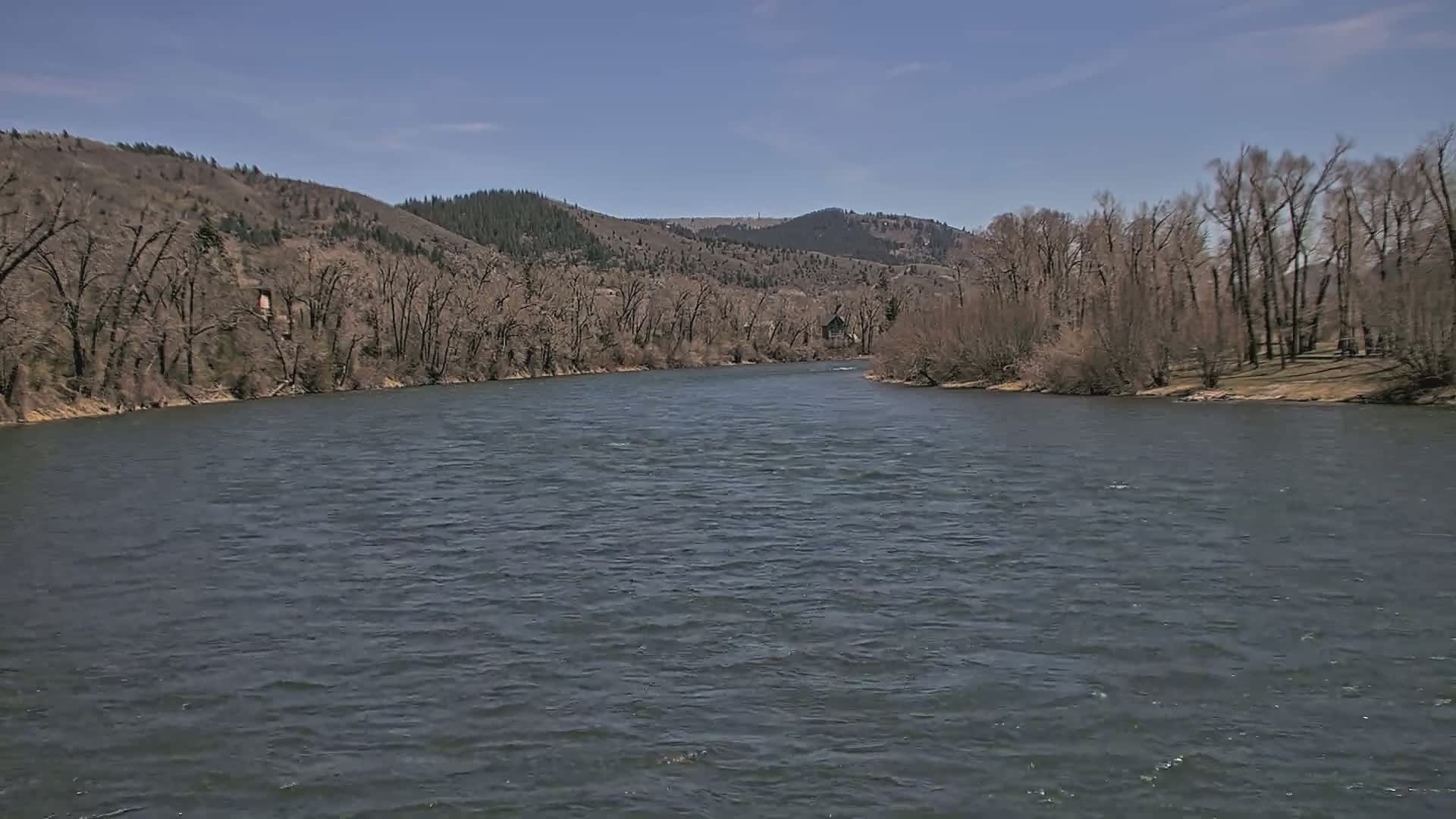 A wide, dark-grey river flows gently under a clear blue sky past bare, brown trees and hills.