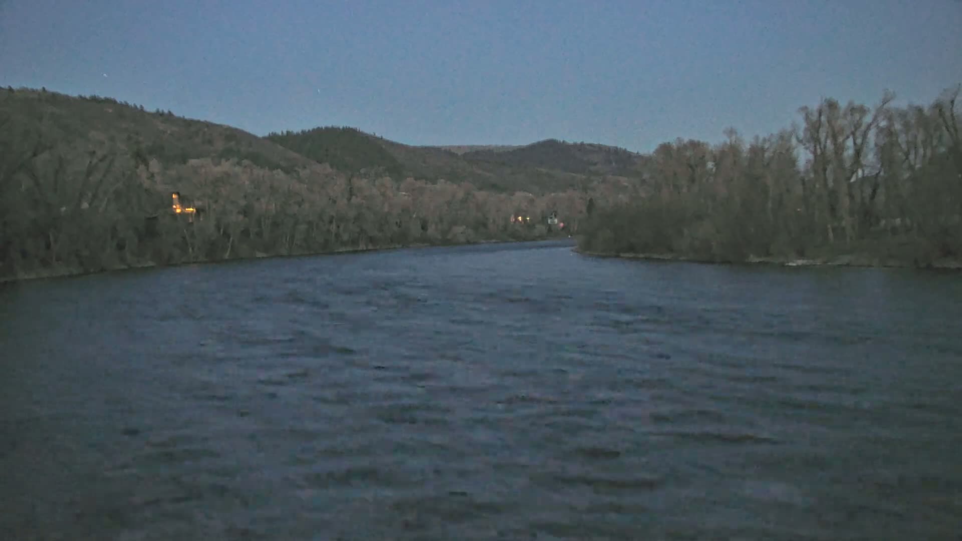 A wide, dark river flows under a twilight sky past dark, leafless trees and hills.