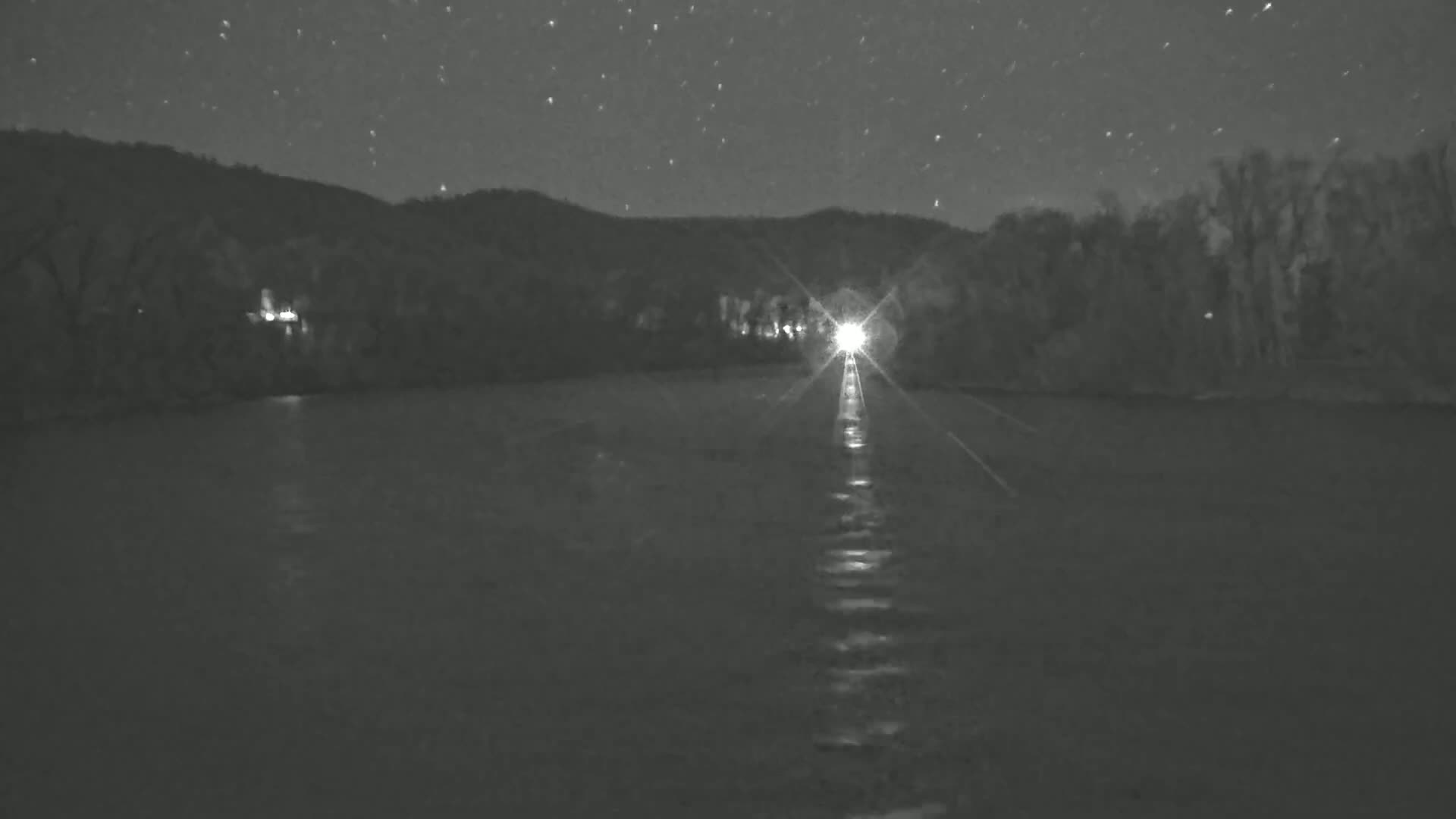 A bright light shines across a dark, still body of water toward a treeline under a starry night sky.