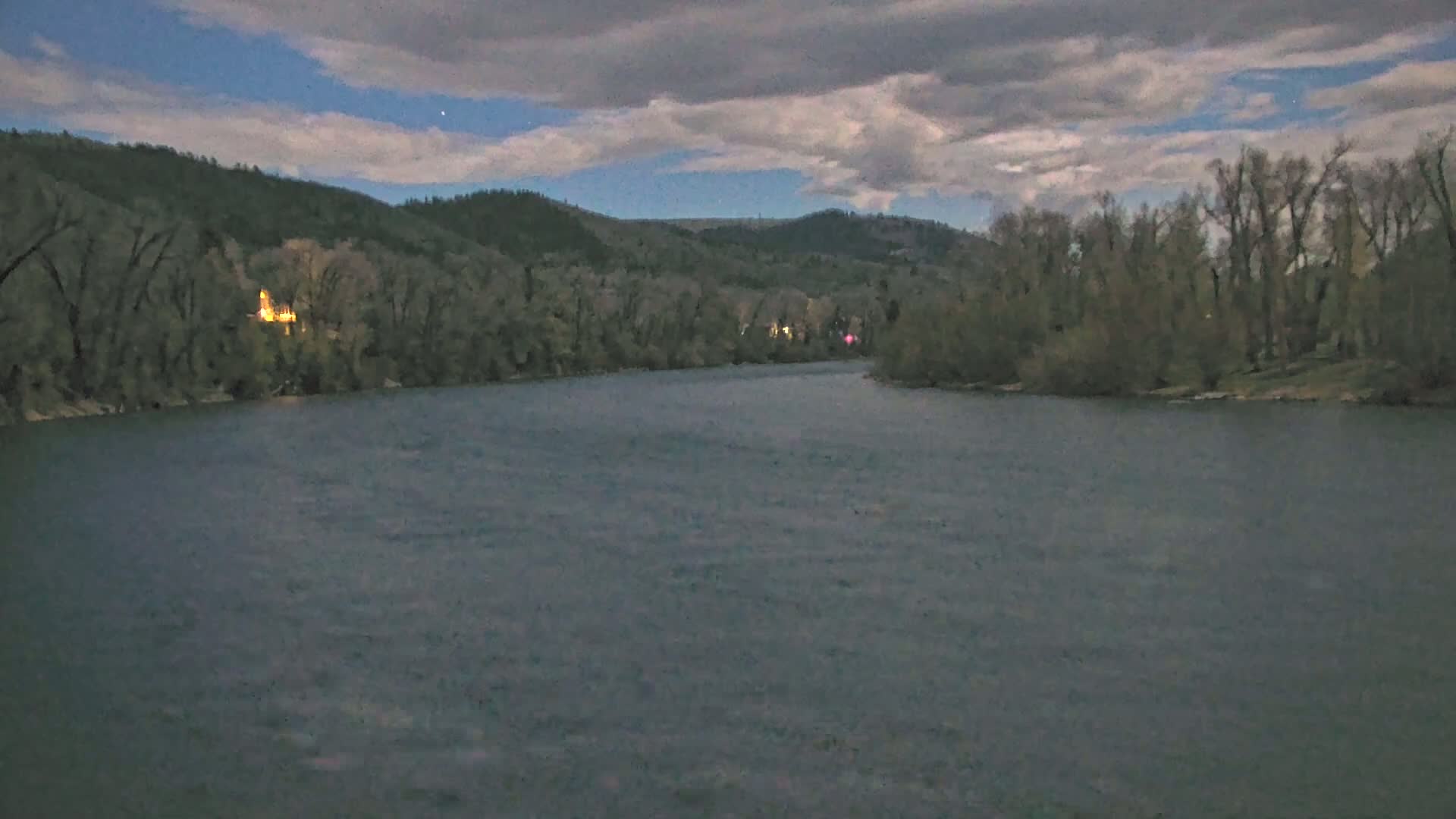 A wide, dark river flows under a cloudy twilight sky, past tree-lined banks.