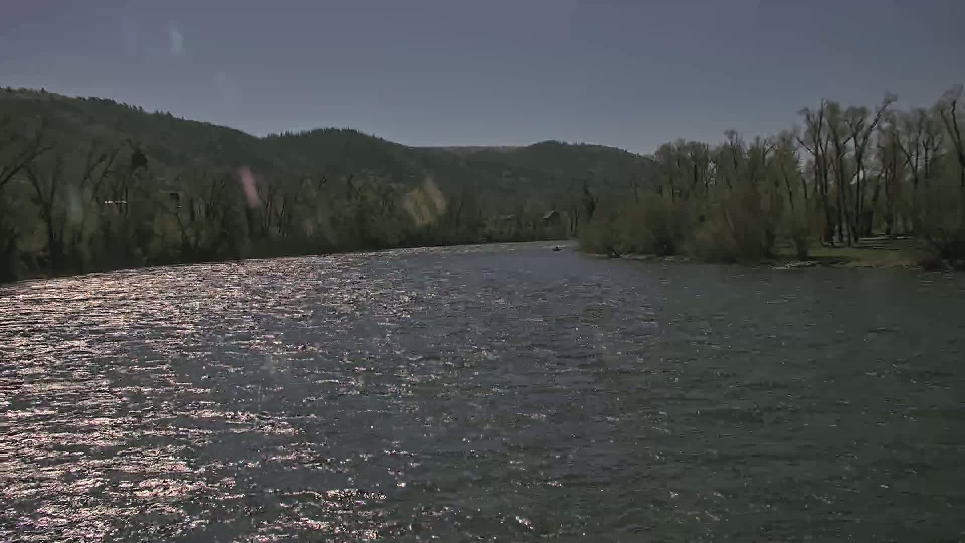 A wide, gently flowing river reflects sunlight on its surface, surrounded by dark green hills and trees under a clear, bright sky.