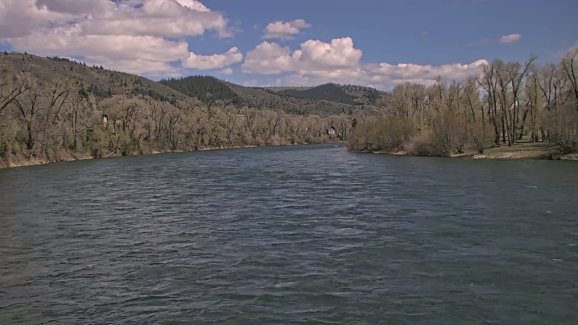 A wide, dark river flows under a partly cloudy blue sky past bare trees and hills.