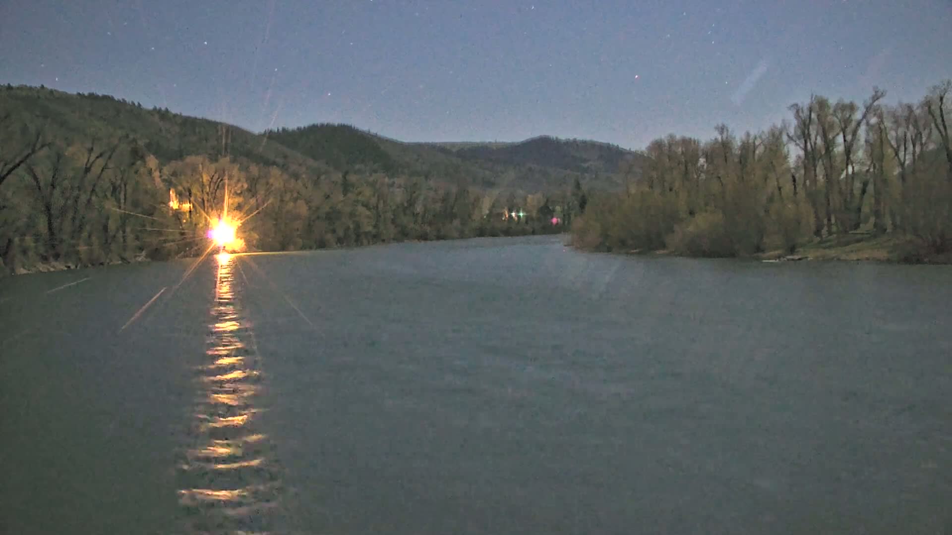 A bright light shines from a shoreline across a dark, calm river under a starry night sky.