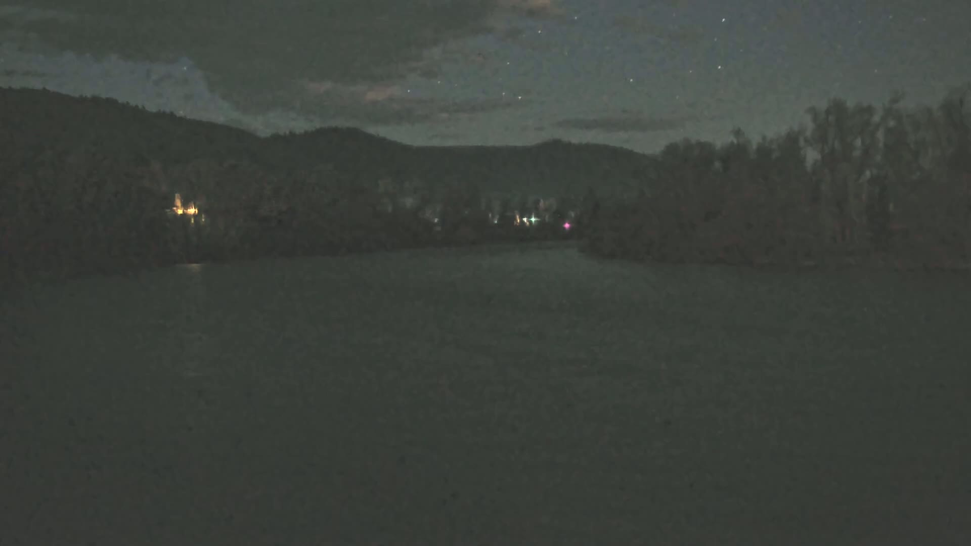 Under a starry, nighttime sky, a dark body of water reflects the faint lights of a distant shoreline and hills.