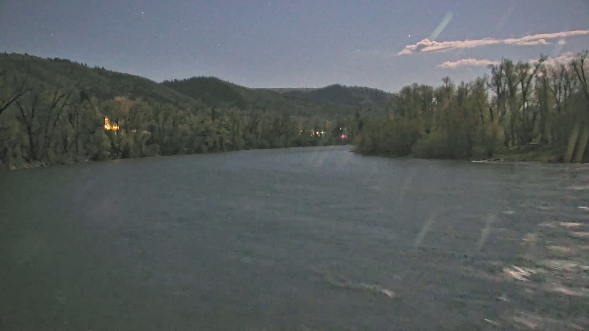 A dark, calm river flows under a twilight sky past tree-lined banks and distant hills.