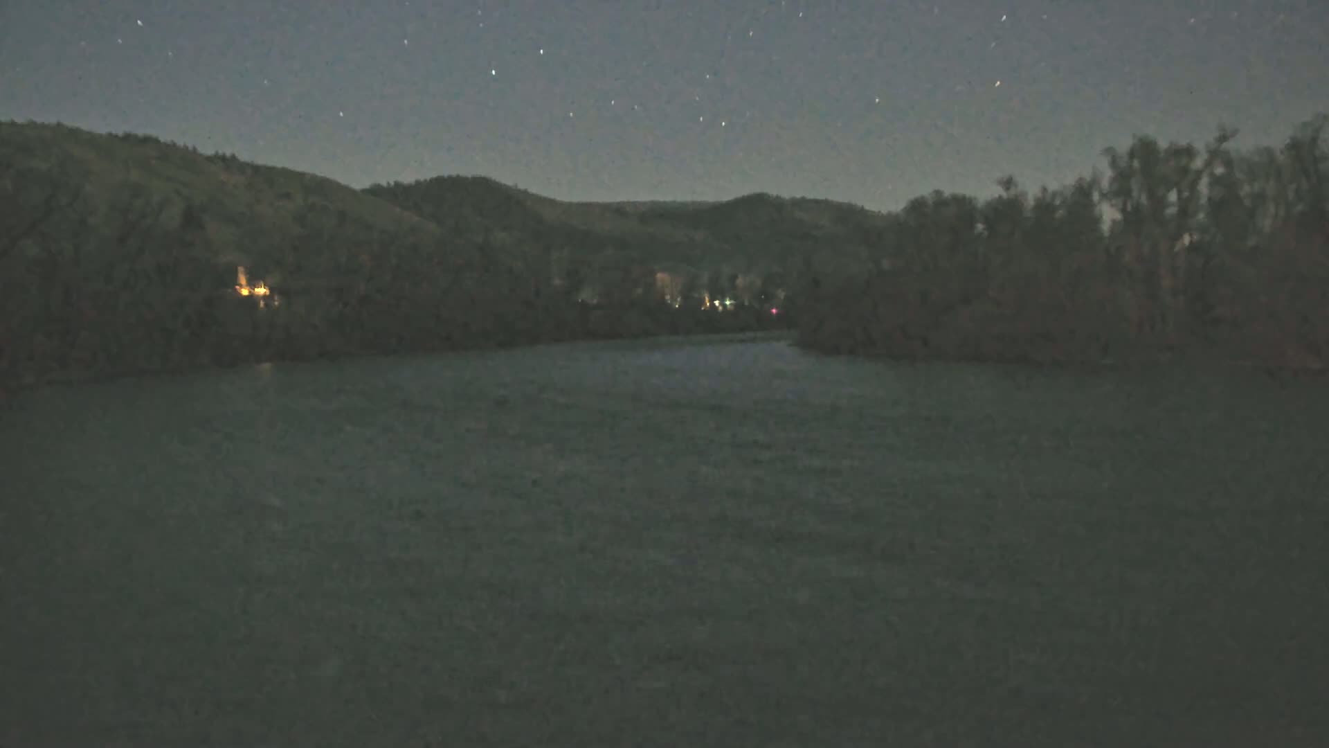 A dark river flows under a starry night sky between hills covered in dark trees.