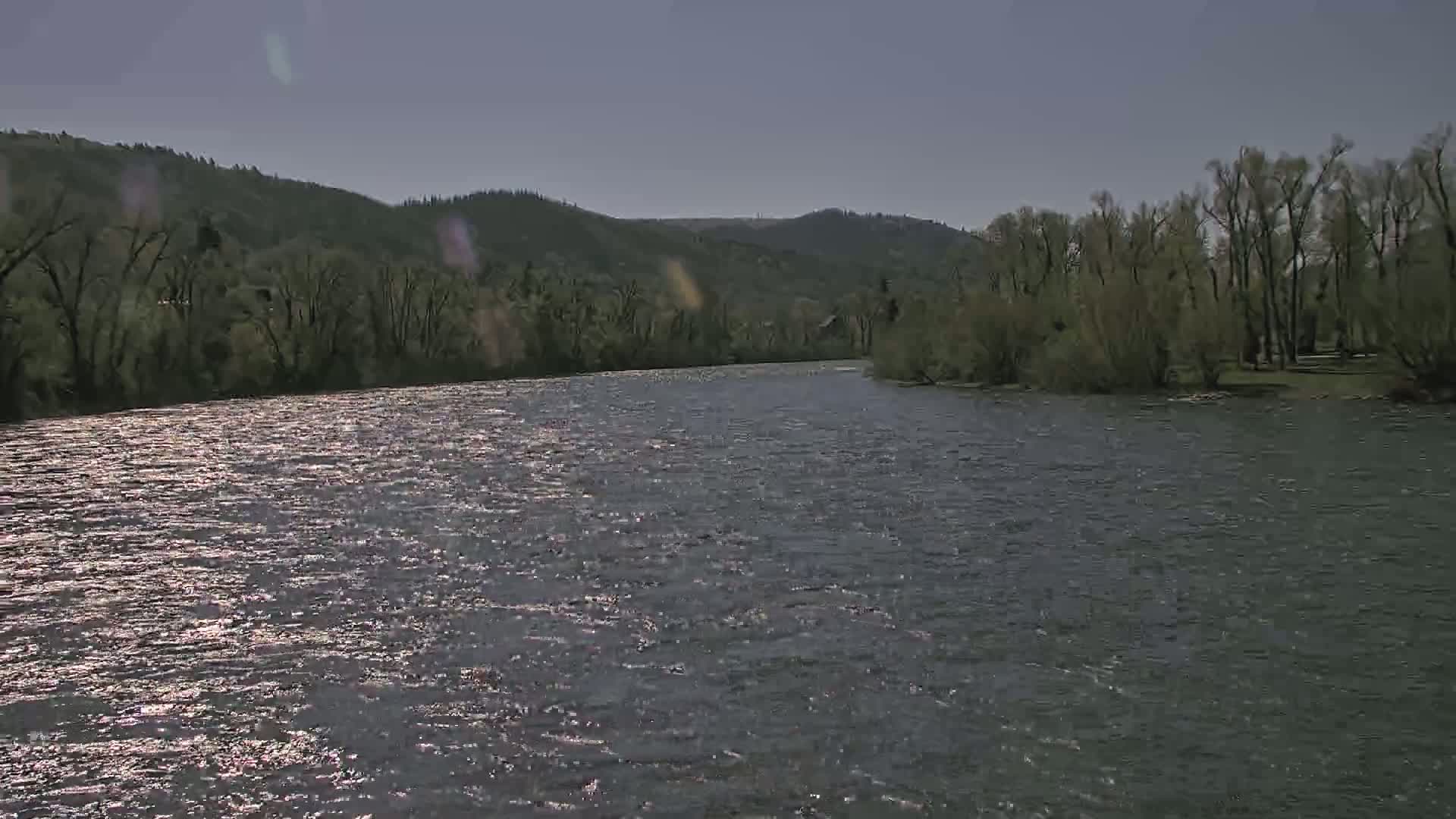A gently flowing river reflects sunlight under a mostly clear sky, bordered by verdant hillsides and trees.