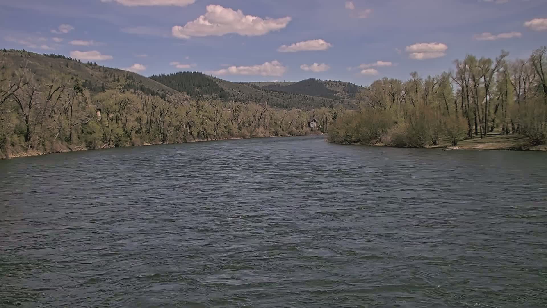 A wide, dark river flows under a partly cloudy blue sky past banks lined with leafing trees and hills in the background.