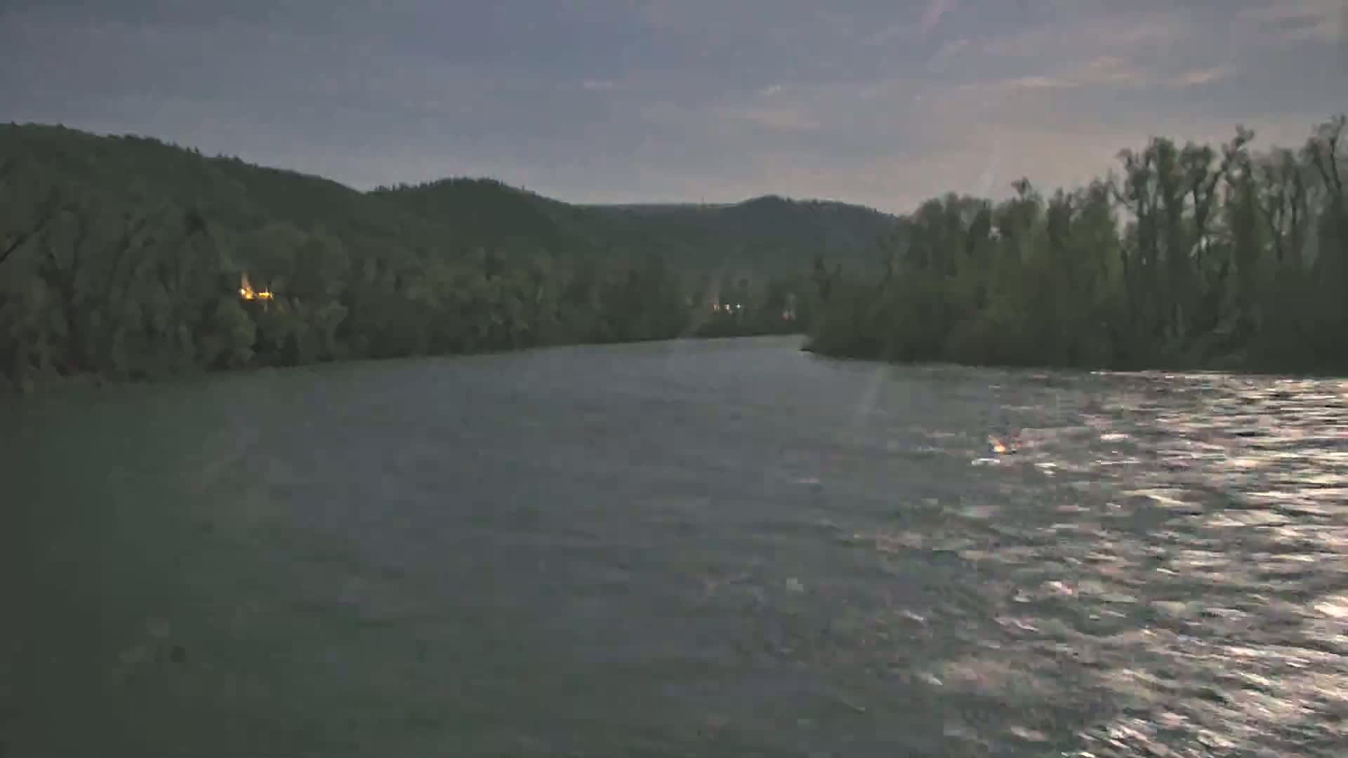 A dark river flows under a twilight sky past tree-lined banks and hills.