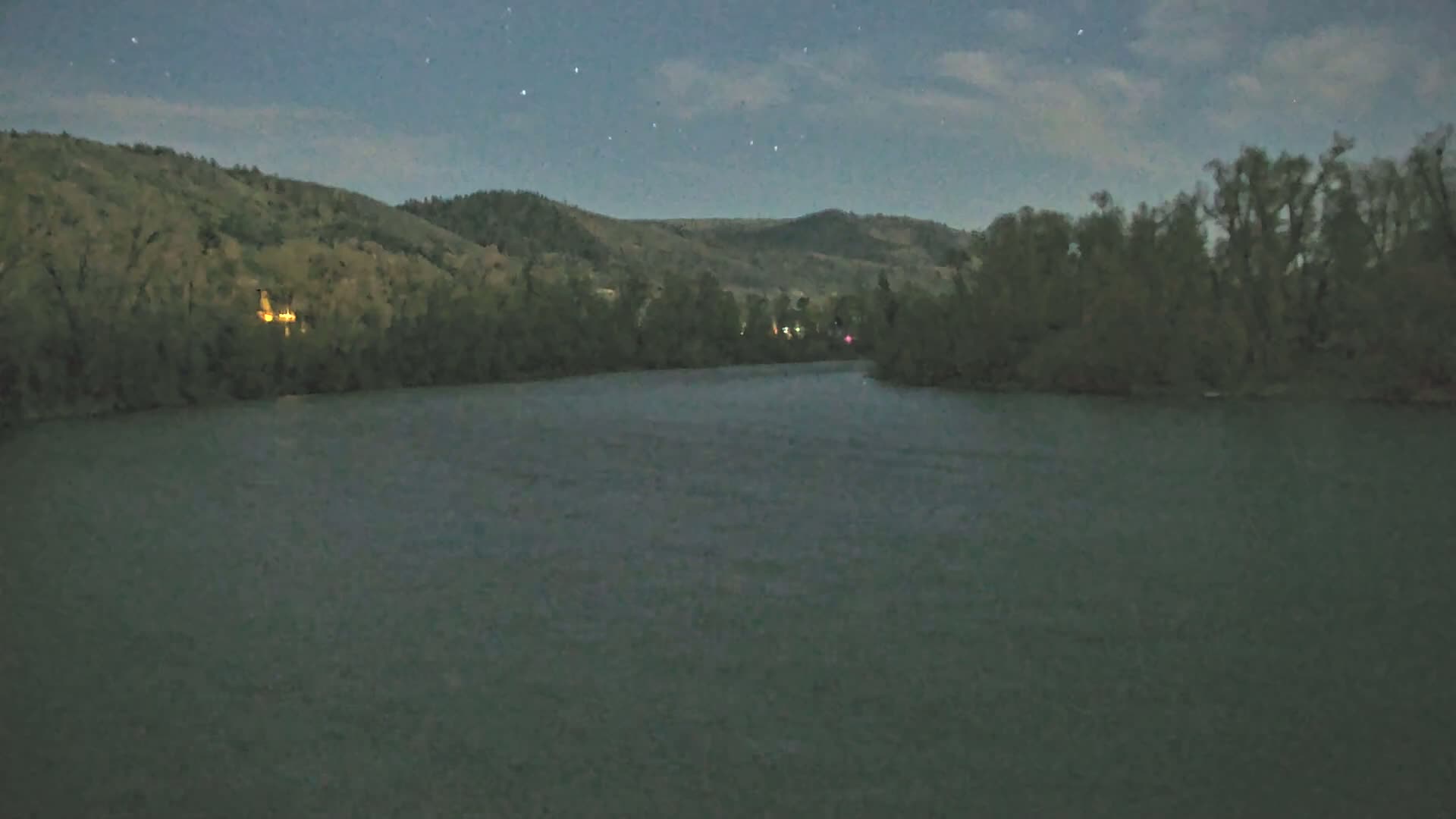 A wide, dark river flows under a night sky dotted with stars, past tree-lined hills.