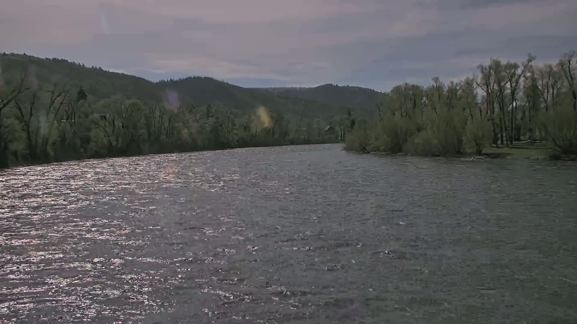 A wide, gently rippling river flows under an overcast sky past wooded banks and hills.