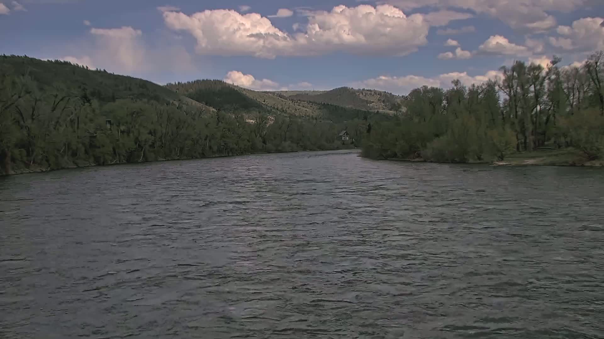 A wide, gently flowing river curves past a lush green shoreline of trees and hills under a partly cloudy sky.
