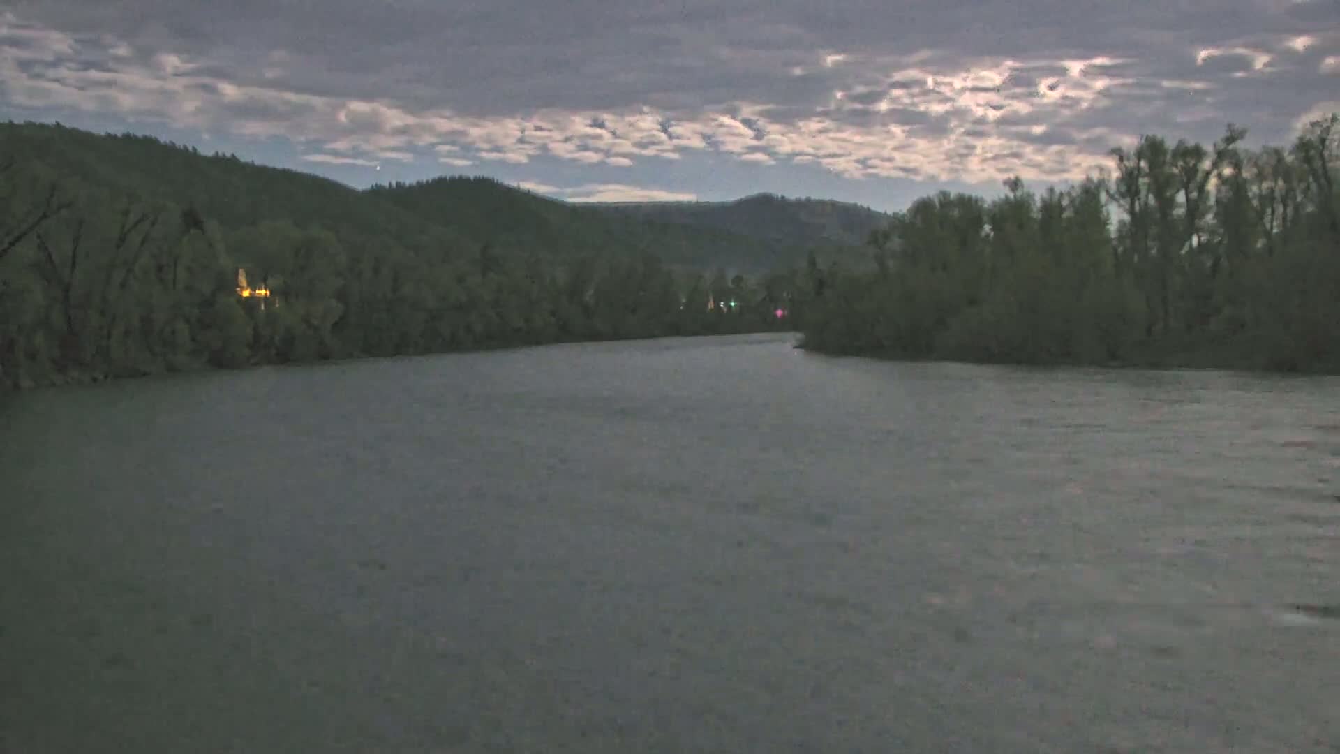 A wide, dark river flows under a cloudy, moonlit sky, past forested hillsides.
