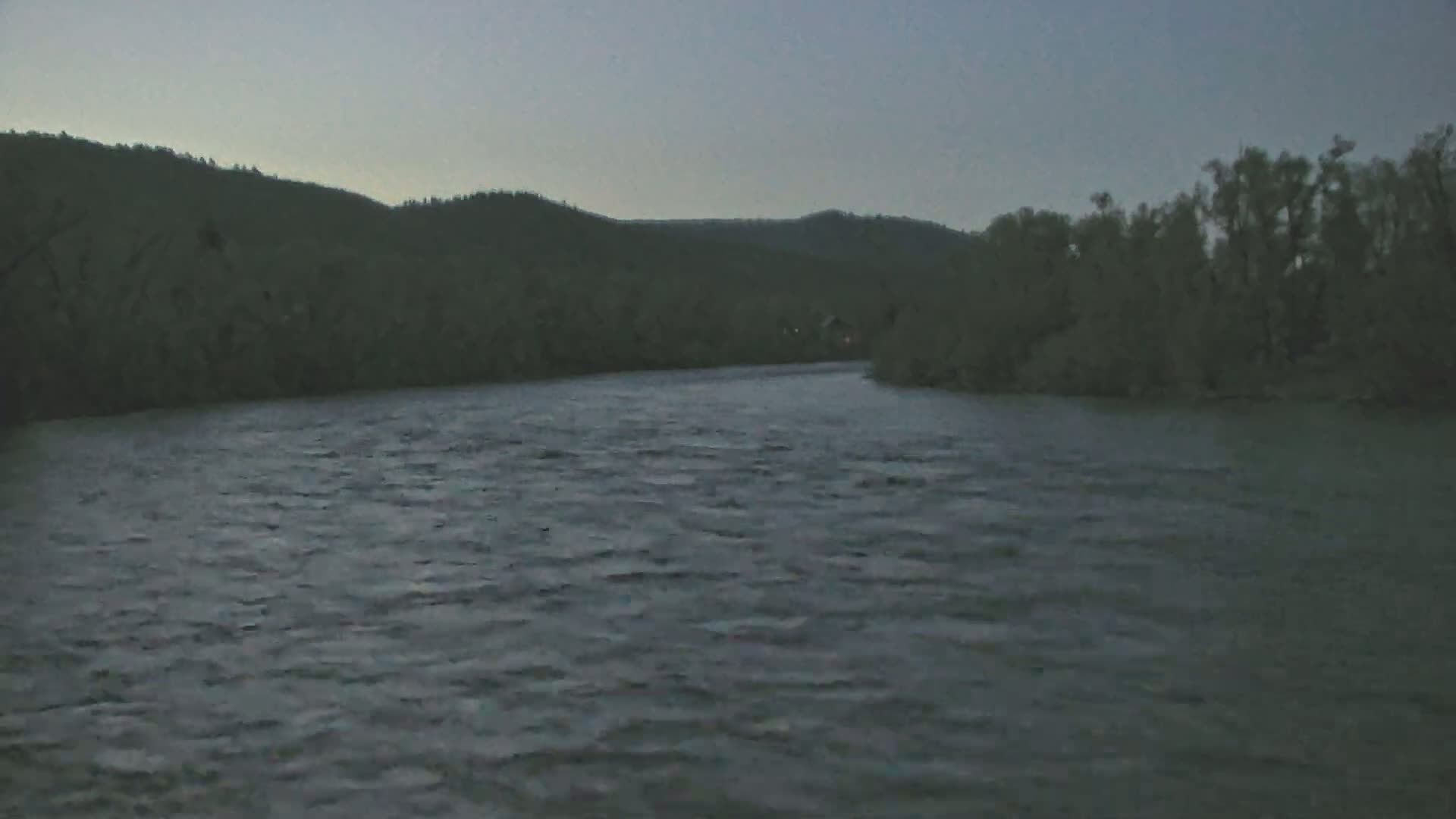 A dark, somewhat choppy river flows beneath a dusky sky, past tree-lined banks and hills in the distance.