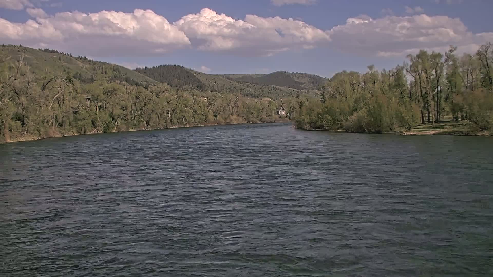 A wide, dark river flows under a partly cloudy sky past tree-lined banks and hills.