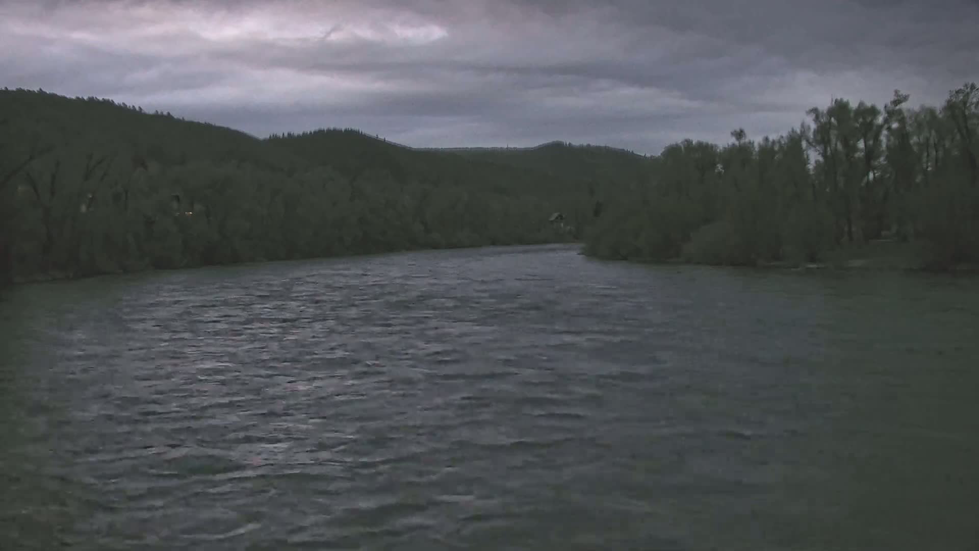 A wide, dark river flows under a cloudy, overcast sky toward forested hills.