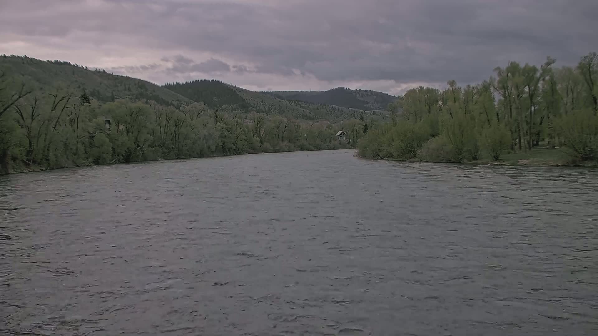 A wide, gray river flows under an overcast sky, bordered by lush green trees and hills in the background.