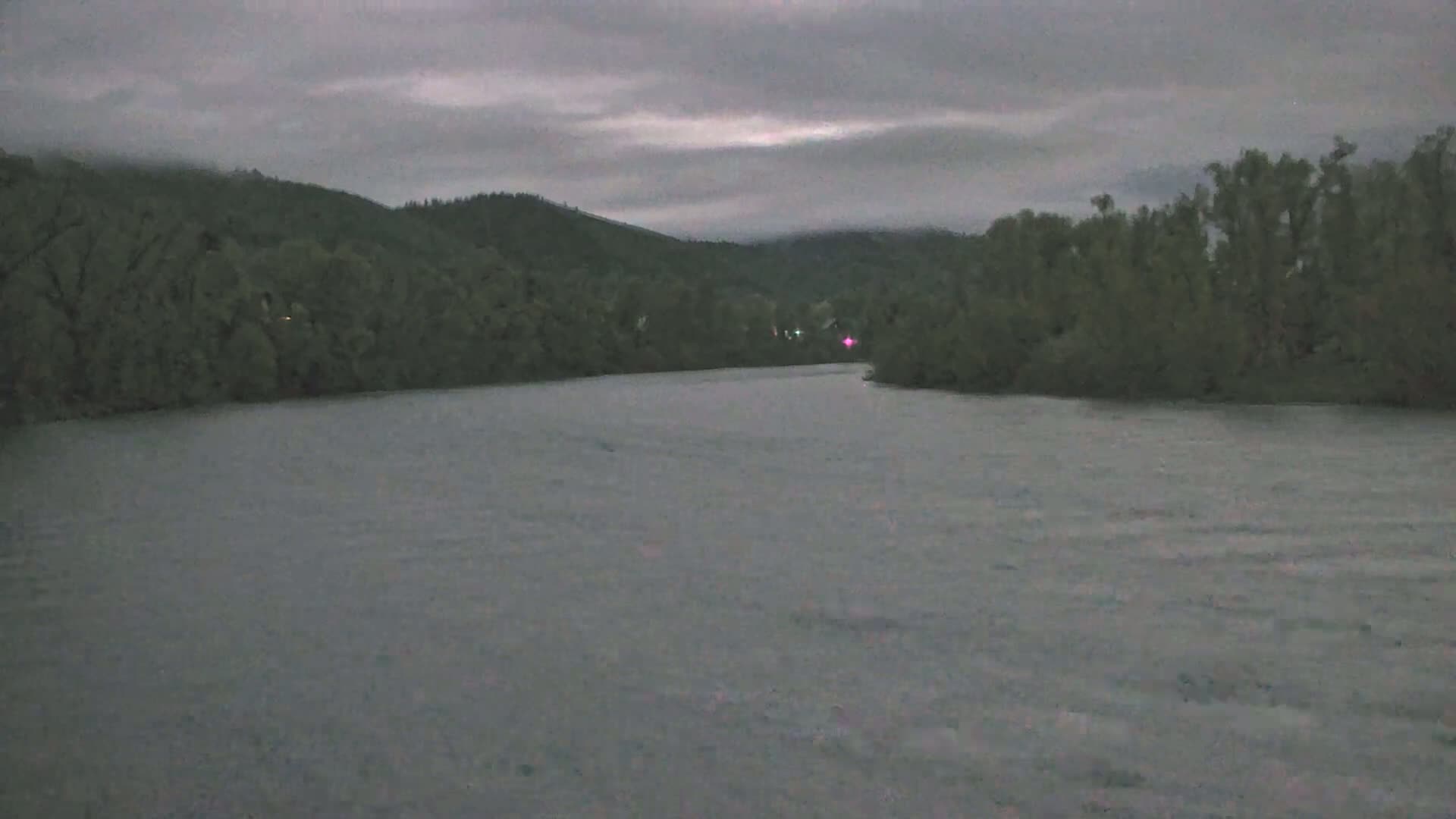 A wide, grey river flows under an overcast sky, past dark green, tree-lined banks and hills.