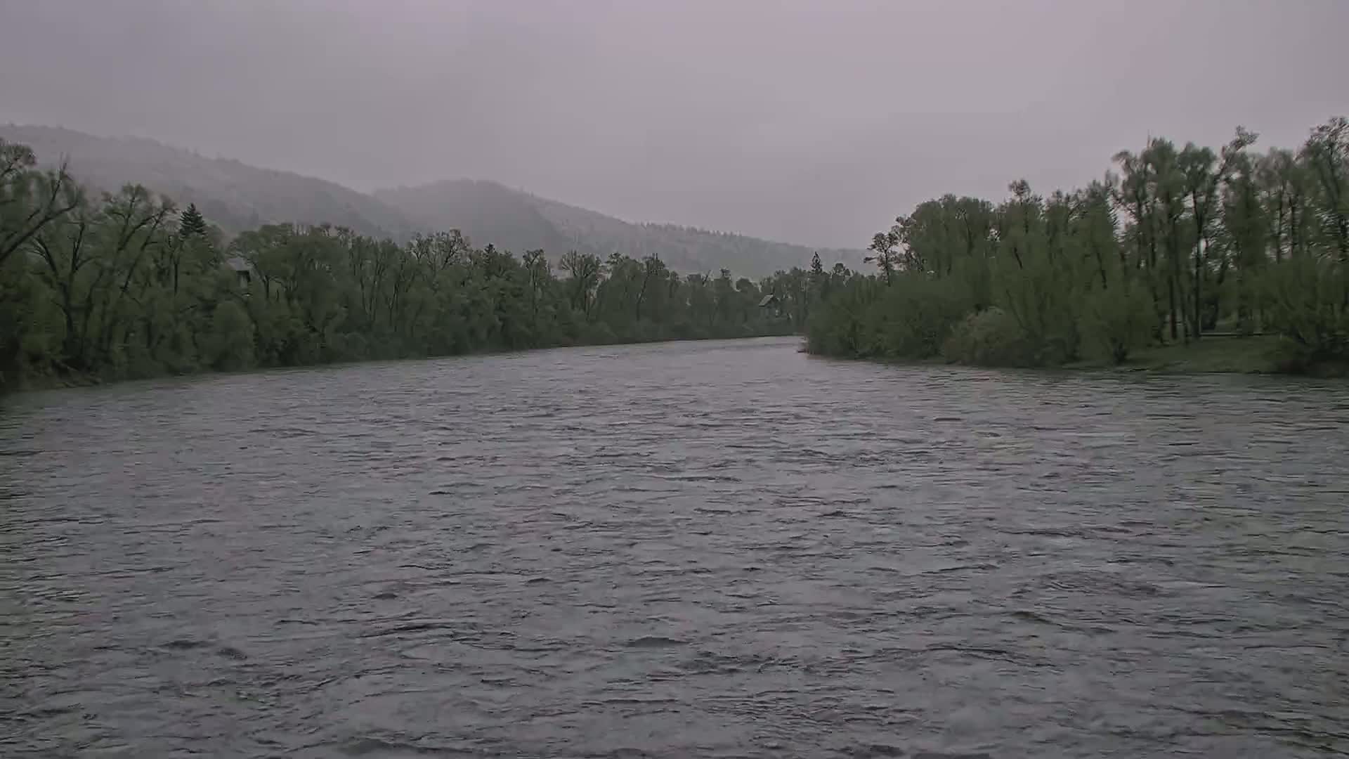 A wide, gray river flows under an overcast sky past verdant banks lined with trees and distant hills.