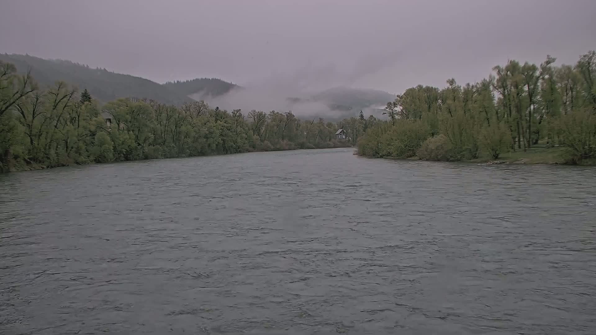 A wide, gray river flows under a cloudy sky past a line of green trees and a distant, fog-shrouded mountain range.